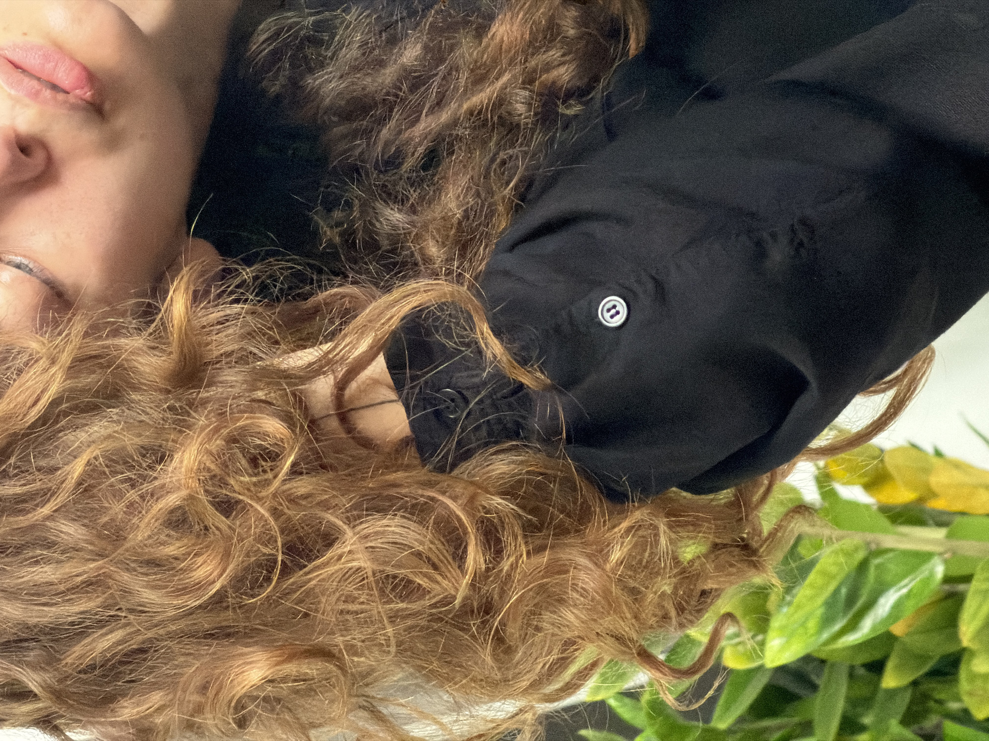 The image shows a close-up, slightly rotated view of a person lying down. The person's face, including the lips, nose, and one eye, is visible. The person has curly, golden-brown hair. A black, buttoned sleeve is positioned near the person's head. A green, leafy plant is in the bottom right corner of the image. The location is not discernible from the information provided.