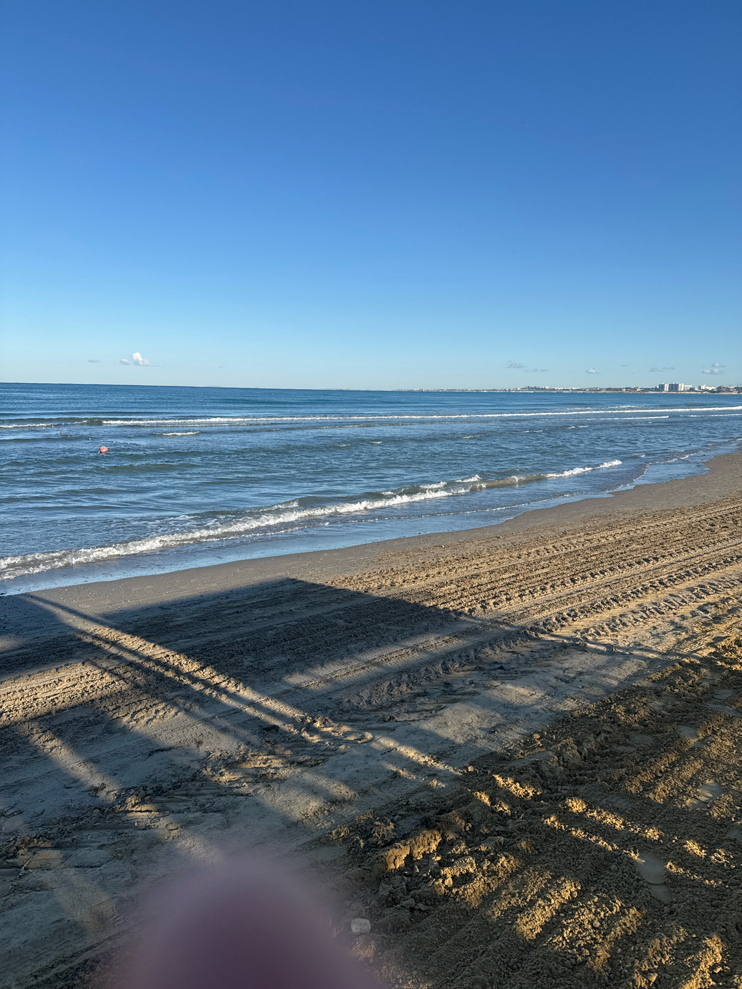 The image depicts a wide-angle view of a sandy beach under a clear blue sky. The foreground is dominated by wet sand with tire tracks and shadows cast from an unseen object, possibly a structure or vegetation. A blurry, pinkish object, likely a person's finger or hand, is partially visible in the lower left corner of the frame.
The middle ground features gentle waves rolling onto the shore, with white foam cresting on the water's surface. A red buoy is visible in the sea, approximately one-third of the way from the left edge of the image. The water is a deep blue, transitioning to a lighter hue near the shore.
In the background, a thin line of buildings and structures can be seen along the distant coastline, suggesting the presence of a city. A few small clouds are scattered across the horizon. The scene is consistent with a daytime coastal environment, possibly during a calm weather period.
There is no specific information within the image to identify the location as Anapa, Russia.