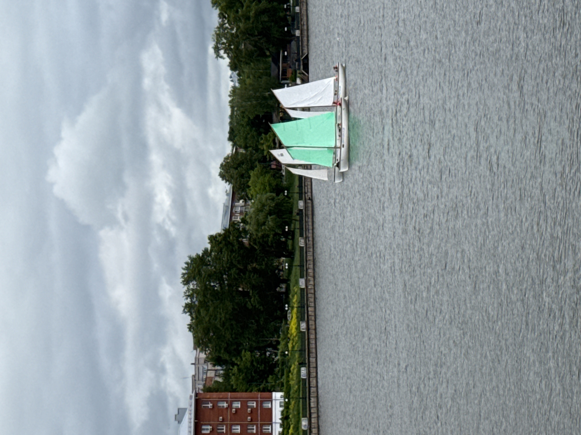 The image displays a daytime scene in Votkinsk, Russia, featuring a body of water with a developed shoreline under a heavily overcast sky. In the midground, three small sailboats are positioned on the water. The central sailboat has a prominent green main sail, while the boat immediately to its left and the boat to its right both have white sails. Small human figures are visible within the hulls of these boats.
The far bank of the water is lined with a structured embankment, above which a strip of green foliage interspersed with yellow flowers is present. A paved pathway runs parallel to the water, illuminated by several dark-colored lampposts, each equipped with multiple light sources, spaced at regular intervals. A single individual is discernible walking along this path on the far bank.
The background consists of a dense area of green trees, suggesting a park or wooded zone. To the left, a multi-story building constructed of red brick with white window frames is visible, featuring numerous rectangular windows and external fixtures on its facade. Other urban structures are partially obscured by the trees and further in the background. The sky is completely covered by thick, grey clouds, indicating an overcast weather condition.