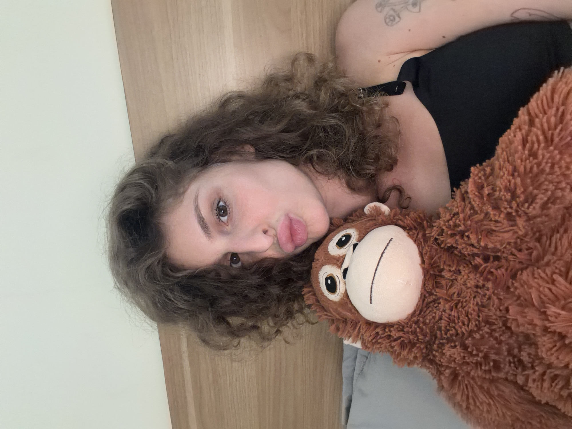 A young woman with curly brown hair and a black tank top is posing for a selfie in a bed. She is holding a brown stuffed monkey and puckering her lips. Her right arm is visible, with a tattoo on her upper arm. The background shows a light-colored wall and a wooden headboard. The scene appears to be indoors, in a bedroom, potentially in a hotel or residential setting in North Kuta, Indonesia.