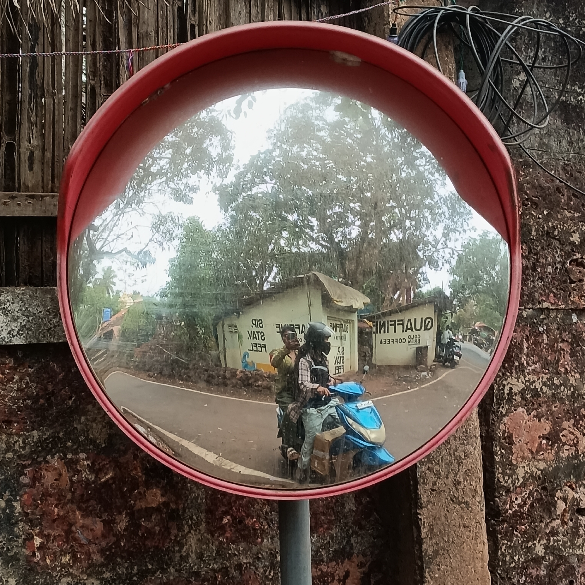 A convex safety mirror with a red frame is mounted on a gray metal pole. The mirror reflects a street scene in Assagao, India. The reflection shows two people on a blue motor scooter driving toward the mirror's location. A building with the word "QUAFFINO" painted in reverse on its side is visible behind the scooter. A person in the reflection is holding a cell phone. The background includes trees and other vegetation. The mirror is situated against a weathered wall. Electrical cables are visible above the mirror to the right. A fence made of thin sticks is above the mirror on the left.