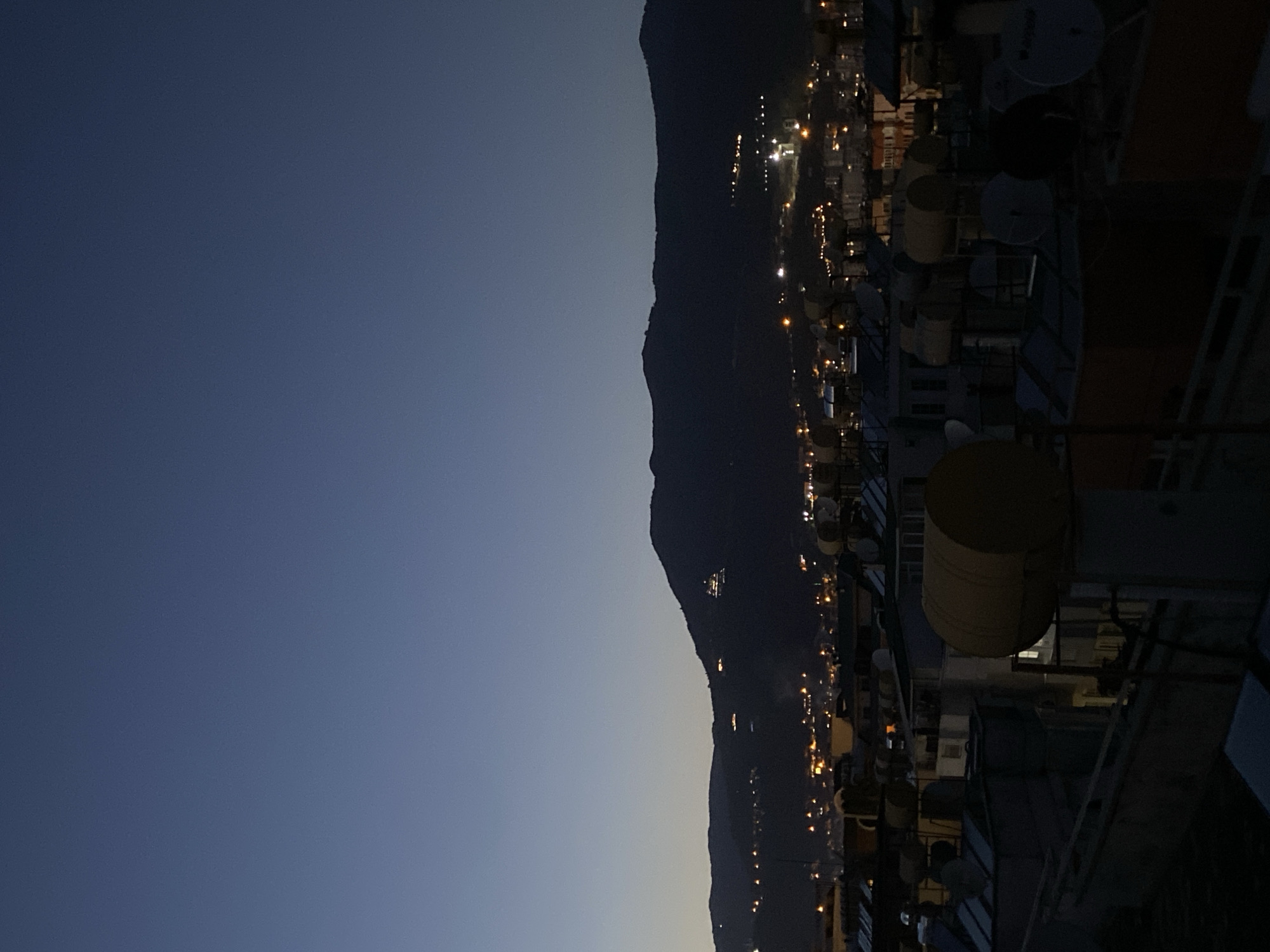 The image captures an expansive, elevated view of a city nestled at the base of a dark mountain range during twilight. The primary subjects include the mountainous landscape, the urban area with its myriad lights, and various rooftop installations in the foreground.
In the foreground, on the right side of the frame, are elements of a building's rooftop. Visible objects include several large, cylindrical tanks (likely water heaters or storage) in shades of yellow and beige, multiple satellite dishes, and what appear to be air conditioning units and other utility structures. Parts of railings or structural supports for a building are also discernible, suggesting the photo was taken from a high vantage point, possibly another building's rooftop or balcony.
The midground is dominated by the silhouette of a formidable mountain range, rising steeply from the city. Its dark, undulating peaks contrast with the fading light of the sky. Scattered across the mountain slopes and extending into the valley below are countless small, bright yellow and orange artificial lights, indicating homes, streets, and other urban infrastructure. These lights create a warm glow against the darkening landscape.
The sky, occupying the upper left portion of the image, displays a gradient of twilight colors, transitioning from a deeper, muted blue at the top to a lighter, almost greyish-blue closer to the horizon. The absence of stars suggests it's either dusk just after sunset or dawn before sunrise.
No specific activity or event is taking place within the frame, but the scene conveys the quiet transition from day to night or night to day in a bustling city. The weather appears clear, with no clouds visible. The location, given the context, is Alanya, Turkey, identifiable by the characteristic mountainous coastal setting with urban development extending up the slopes. There is no visible text in the image.