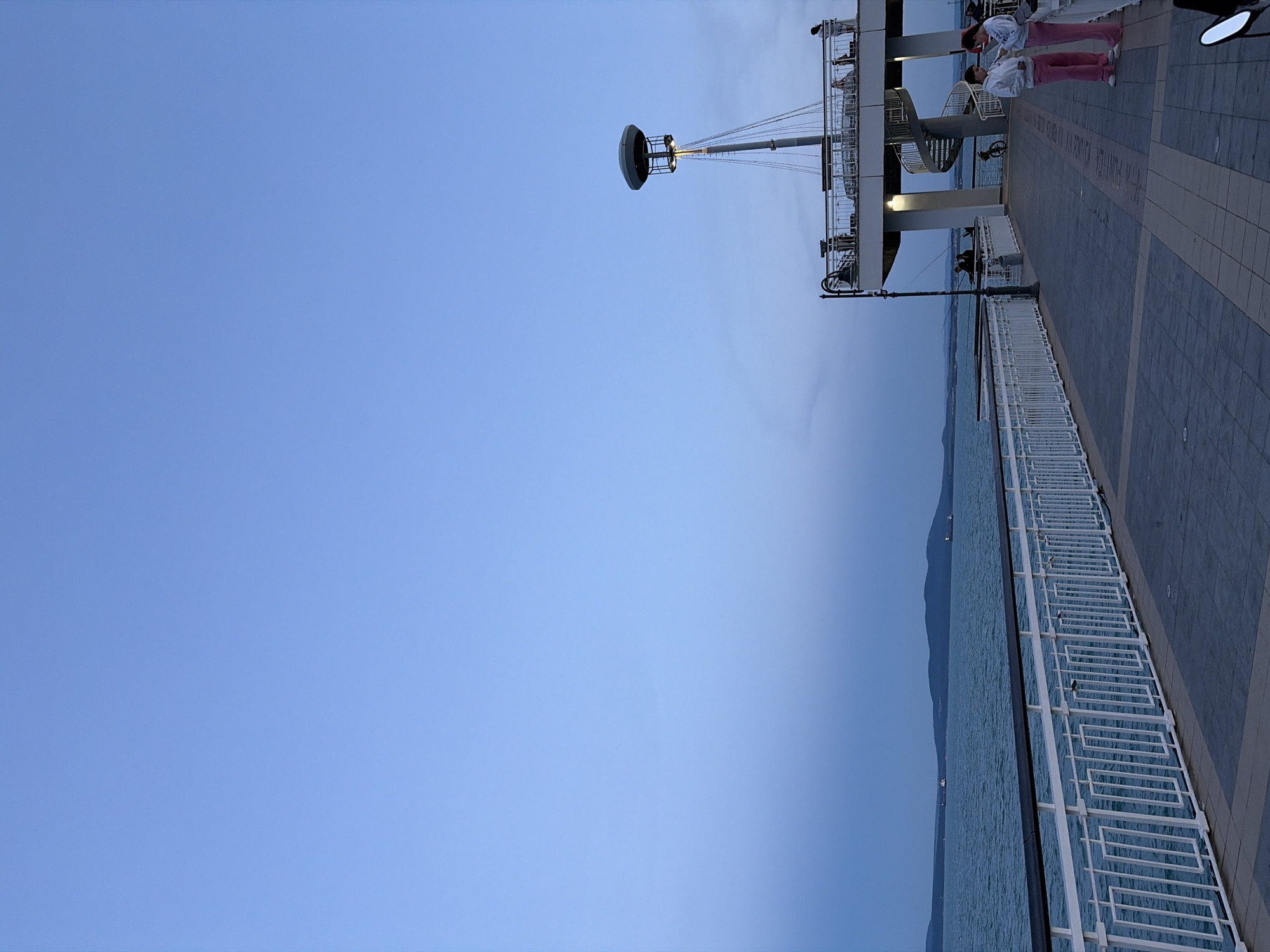 The image captures a serene dusk scene along the Burgas Sea Garden's alleyway, adjacent to the Black Sea. The expansive sky is a gradient of soft blue, transitioning to a muted grey near the horizon, where distant hills are visible. The tranquil sea, in shades of deep turquoise, stretches out towards the hills. A white, modern railing lines the promenade, separating it from the water.
The central focus on the right is a multi-level structure featuring a prominent light pole with a distinctive mushroom-shaped head. Below it, a white balcony with railings is adorned with several individuals, some standing and observing the view, others in conversation. A spiral staircase descends from this balcony, adding an architectural element. The ground of the promenade is paved with grey and light brown rectangular tiles.
Several people are present. Two young individuals, a male and a female, are standing in the foreground, both wearing pink tracksuits, with the female looking at her phone. Further down the promenade, near a fishing rod propped against the railing, several figures are engaged in fishing. Other individuals are scattered on the upper levels of the structure. The scene is illuminated by the warm glow of the light pole and other ambient lighting, suggesting the early evening. The overall atmosphere is calm and peaceful, characteristic of a leisurely stroll in the park.