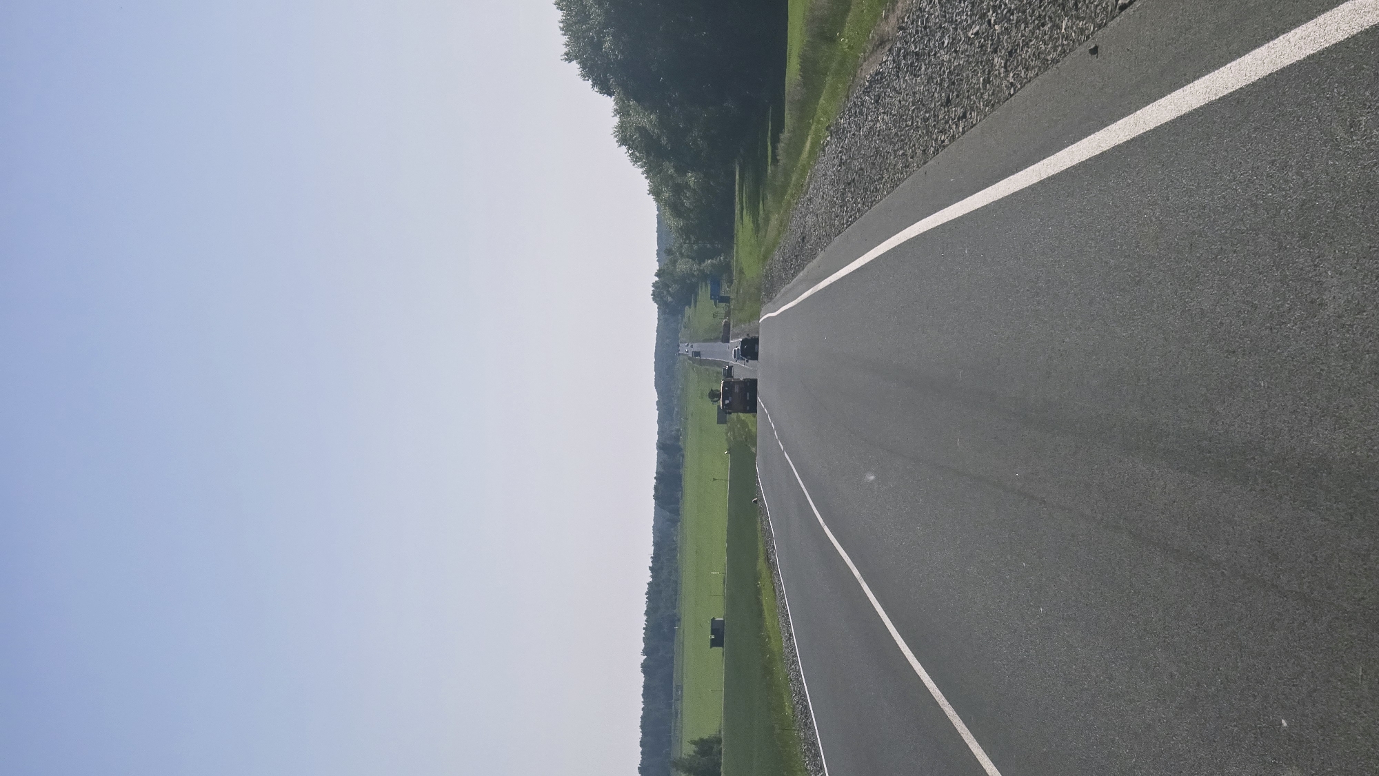 The image depicts a straight, asphalt road with white lane markings, leading towards a distant horizon. The sky is clear and pale blue. On either side of the road, there are green fields and trees. In the distance, several vehicles are visible on the road, including a red truck. The scene appears to be set in a rural area. There are no identifiable buildings or landmarks that would place this image specifically in the city of Актюбинский, Russia. The environment suggests a clear, sunny day.