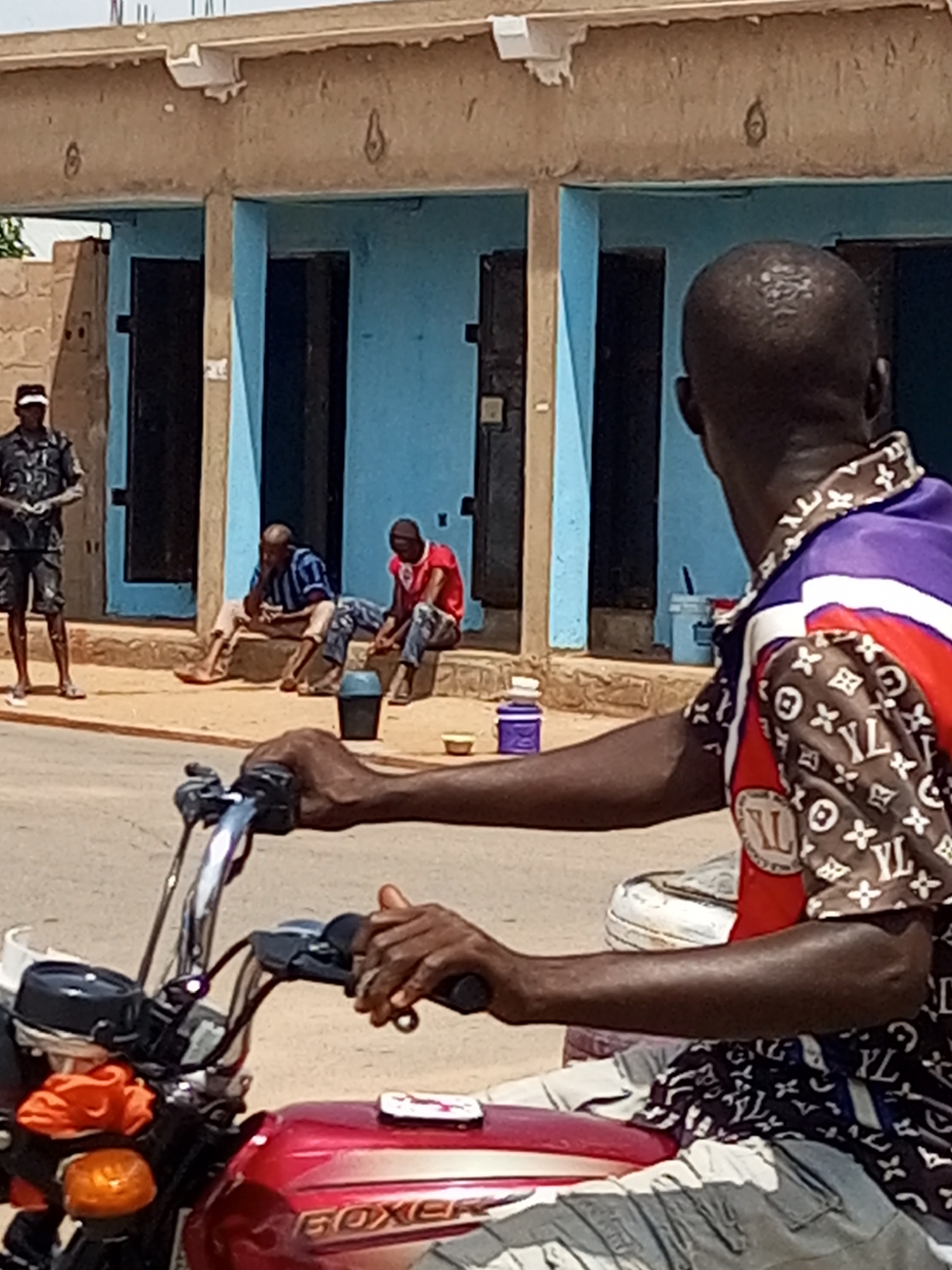 An overcast, daytime scene in Tempure, Nigeria, depicts a street with a light-colored, rough textured pavement in the foreground and a blue building with a beige, concrete overhang in the background. On the right side of the frame, the back of a person on a red motorcycle is visible, wearing a patterned shirt. In the middle ground, two men are seated on a raised concrete ledge in front of the blue building. The man on the left wears a striped blue and white shirt and light-colored pants, and the man on the right wears a red t-shirt and dark pants. To the left of the men, a black bucket and a small yellow bowl are on the ground. To the far left of the frame, a person in a dark uniform and hat stands on the pavement. The building has several recessed doorways with dark doors.