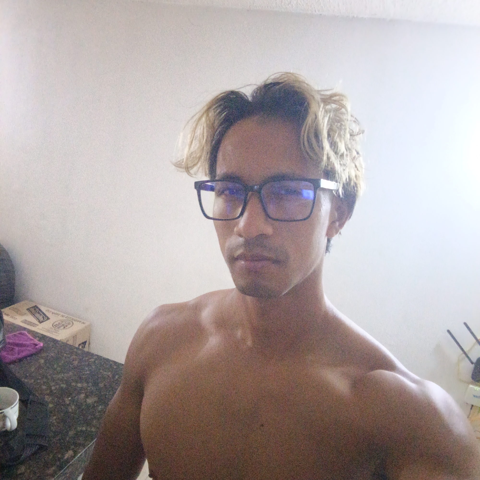 A shirtless man with blonde hair and glasses is taking a selfie in a room. He is looking directly at the camera. In the background, there is a wall and a counter with a box and a cup on it.