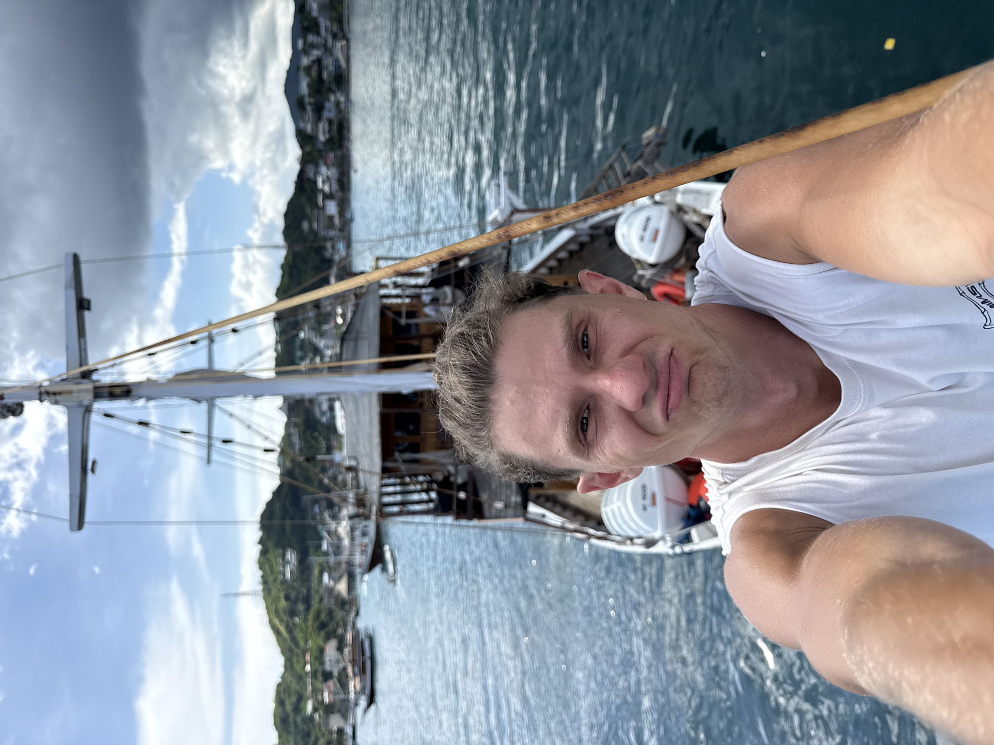 A man in a white tank top is taking a selfie on a sailboat. The man is facing the camera, and his right arm is visible on the right side of the frame, presumably holding the phone. Behind him is the sailboat's mast and rigging, and in the background is a body of water with land visible in the distance. The city of Labuan Bajo, Indonesia is likely the location. The water is blue with some reflections. The sky is partially cloudy.