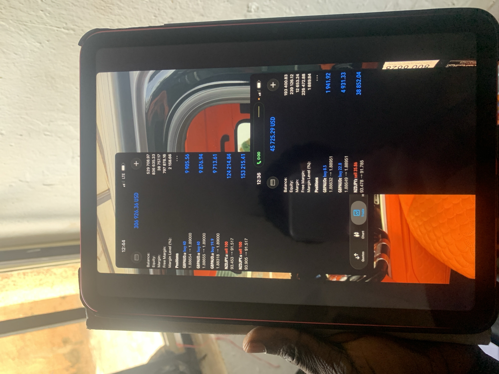A person is holding a tablet displaying two screenshots of a trading platform interface. The screenshots show various financial metrics and open positions, indicating active trading or investment activity. The interface displays information such as balance, equity, margin, free margin, margin level, and specific positions for currency pairs like GBPAUD and NZDJPY. The person's hand is visible in the lower portion of the image, holding the tablet. The background is blurred and indistinct, with a window and vague outlines of objects and the environment. The location in the city is Unknown, Unknown.