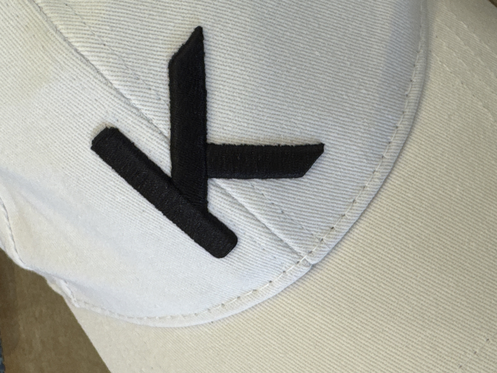 The image shows a close-up, angled view of a light-colored baseball cap with a black embroidered symbol on the front. The symbol resembles a stylized letter "K" or a similar angular character, with a vertical stroke and two horizontal strokes forming an upper "T" shape. The cap is made of fabric with a visible weave pattern. The background is blurred but appears to be a textured surface, possibly a table or furniture. The lighting is bright, creating some highlights on the cap's fabric. There are no people or other discernible objects in the immediate vicinity. The location is unknown.