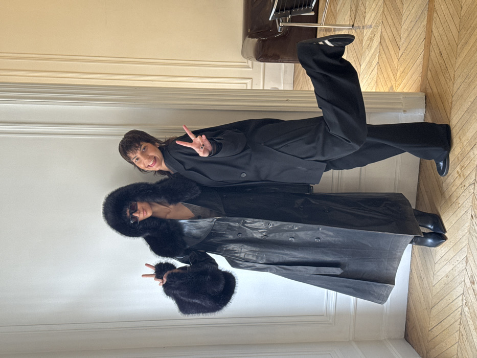 Two adult individuals are posed inside an apartment in Paris, France, indicated by the distinctive herringbone parquet flooring and classical wall moldings.
The individual on the left is tall, wearing a long, dark, double-breasted coat, likely leather, featuring a prominent black fur-trimmed hood and large, matching fur cuffs. Dark sunglasses cover her eyes. She wears dark, pointed-toe ankle boots. Both of her hands are raised, forming peace signs.
The individual on the right, slightly shorter, has dark, layered hair and is dressed in a loose-fitting black blazer, wide-leg black trousers, and dark flat shoes. Her right leg is bent at the knee, with the foot raised, revealing a dark sneaker with a white stripe. She is smiling broadly with an open mouth, facing the camera, and also displays peace signs with both hands. Several rings are visible on her right hand.
The background consists of light-colored walls with decorative molding. A doorway to the right reveals a separate room with matching parquet flooring, a dark brown leather armchair, and a dark-framed chair.