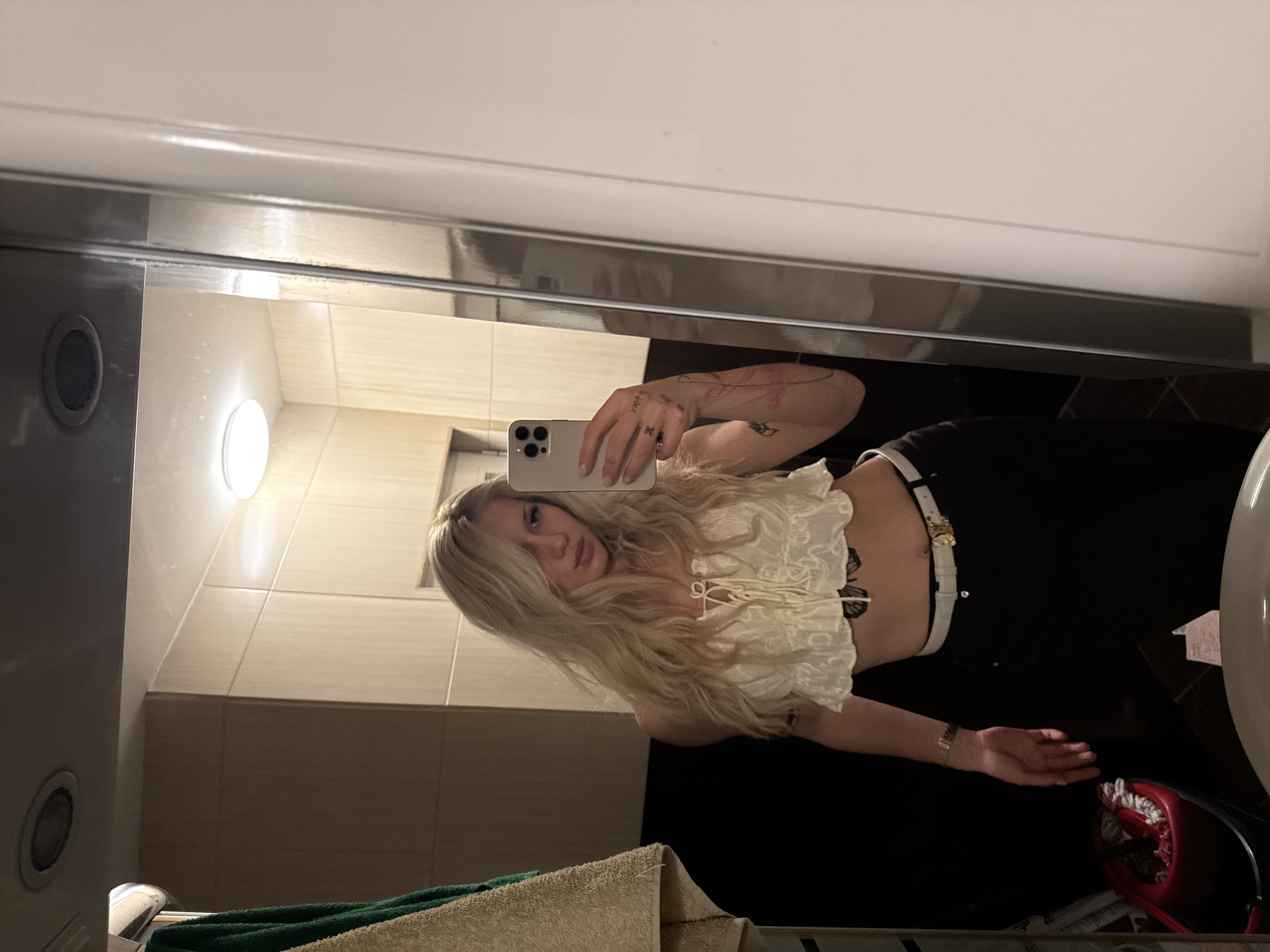 A young adult female with long wavy blonde hair is shown in a mirror selfie, standing in an indoor room in Sofia, Bulgaria. She wears a cream-colored ruffled crop top, black trousers, and a white belt with a gold-toned buckle. Visible tattoos include a large dark butterfly on her abdomen, floral or vine designs on her left forearm, and a small 'X' mark on the back of her left hand. A gold-toned watch is present on her left wrist, and a ring is on her left ring finger. She holds a light-colored smartphone, consistent with an iPhone, in her right hand, angled towards the mirror to capture the image. Her left arm is slightly bent, with the hand open.
The immediate environment, reflected in the mirror, features light beige square-tiled walls and a darker, possibly black, lower wall section. The ceiling exhibits a flush-mounted circular light fixture and a recessed spotlight. To the left of the woman, a towel rack holds two towels: one green and one light beige. Partially visible at the bottom right is a white ceramic fixture, consistent with a sink or toilet bowl. A red object with white components, possibly a container or holder, rests on a dark surface to the lower left. The specific address or establishment within Sofia, Bulgaria, is not visually identifiable.