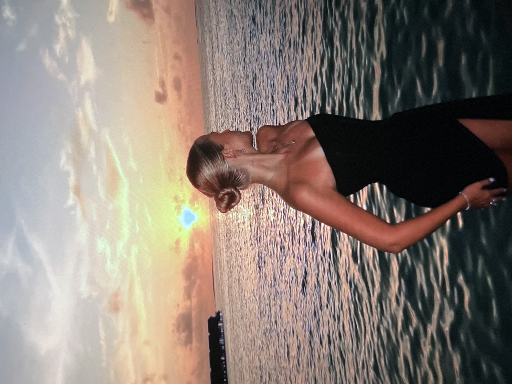 A young woman with blonde hair styled in a bun, wearing a black strapless dress, stands facing away from the camera, looking out at the ocean during sunset. The sun is low on the horizon, casting a warm orange and yellow glow across the sky and reflecting on the water. In the distance, there are overwater bungalows. The scene depicts a serene moment at a coastal resort. The image does not provide any information about the city of Montfermeil, France.