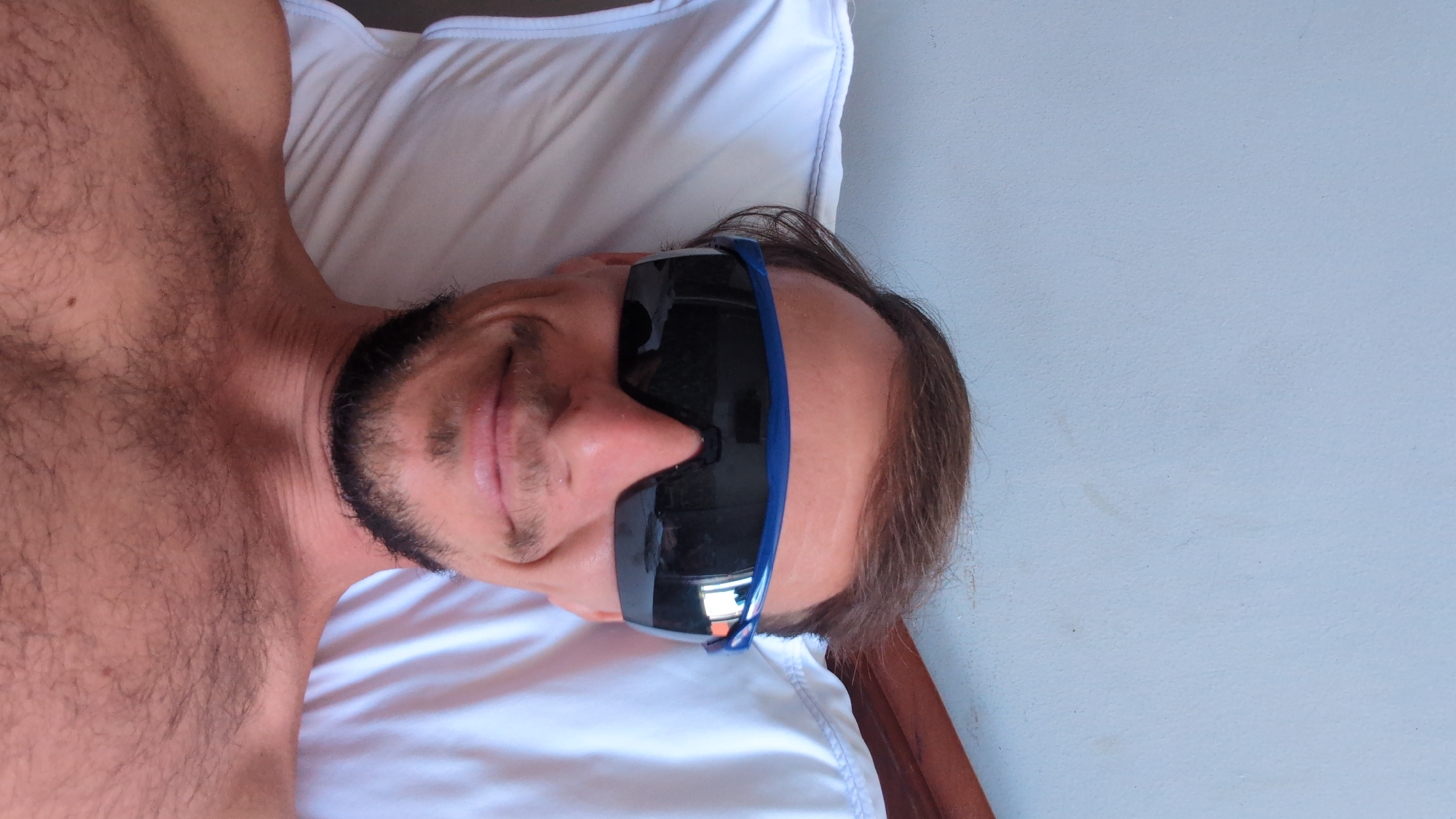 An adult male is depicted in a frontal, close-up, selfie-style image, reclining in an indoor setting within South Quy Nhon Ward, Vietnam. He has short brown hair, a short dark beard and mustache, and is shirtless, revealing a hairy chest and shoulders. A small dark mole is present on his upper right chest. He is wearing blue-framed athletic sunglasses with dark, reflective lenses that obscure his eyes and reflect an indistinct indoor environment, possibly a window. He exhibits a faint closed-mouth smile. His head rests on a white pillow with visible blue stitching along its left edge. A light blue-grey wall serves as the background, with a portion of a dark wooden bed frame visible on the right.