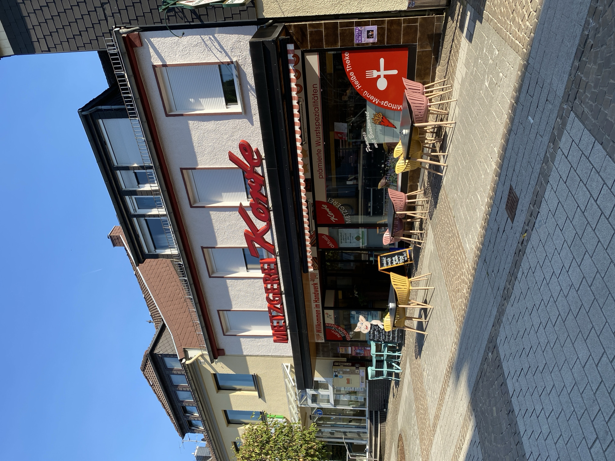 This is a vertical, eye-level, outdoor shot of a butcher shop called "Metzgerei Korte" in Altenkirchen, Germany. The shop has a bright white facade with large display windows and red lettering for its name. The windows advertise "prämierte Wurstspezialitäten" (award-winning sausage specialties) and "Mittags-Menü Heiße Theke" (lunch menu hot counter). Outside the shop, several tables and chairs are set up on a cobblestone sidewalk, suggesting an outdoor seating area. The sun is shining brightly, casting distinct shadows, indicating it is daytime. The sky is clear and blue. A ladder is propped up against the side of the building, possibly for maintenance. The image is a street-level view of a commercial establishment on a sunny day, highlighting its offerings and inviting atmosphere.