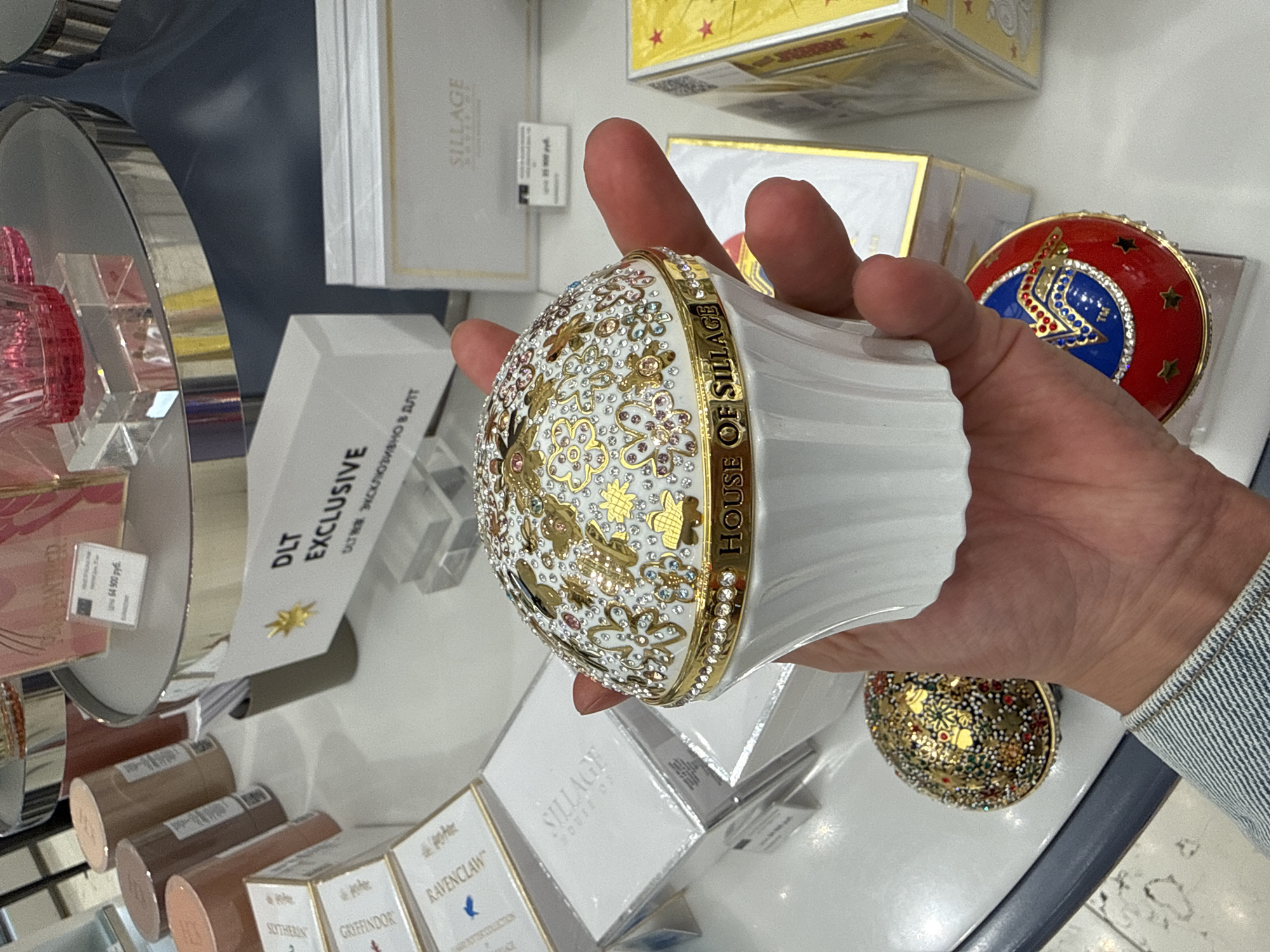 In Saint Petersburg, Russia, at a retail display, a person's hand holds a white, ornate, cupcake-shaped decorative object adorned with gold embellishments and small colorful rhinestones. The object has the inscription "HOUSE OF SILLAGE" around its base. The background reveals other merchandise, including several white boxes with gold lettering, one box labeled "Sillages," and a gold-colored, bejeweled sphere. Another decorative egg-shaped object, red and blue with star motifs and what appears to be a shield emblem, is visible near the bottom right. A white sign with the text "DLT EXCLUSIVE" is also present. The lighting suggests an indoor retail environment.