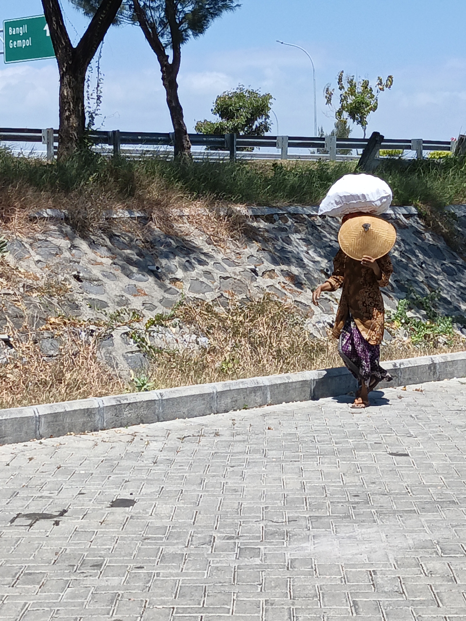 A person walks on a paved walkway beside a road, carrying a bundle on their head. The person is wearing a brown patterned top and a purple patterned skirt. They are also wearing a wide-brimmed hat. The walkway is made of grey interlocking pavers. To the left of the walkway is a grey concrete curb, and beyond that, a slope covered in rocks and dry grass. In the background, there are trees, a highway guardrail, and a blue sky with some clouds. A green road sign indicating "Bangil Gempol 1" is visible in the upper left corner. The scene appears to be in a semi-rural or suburban area.