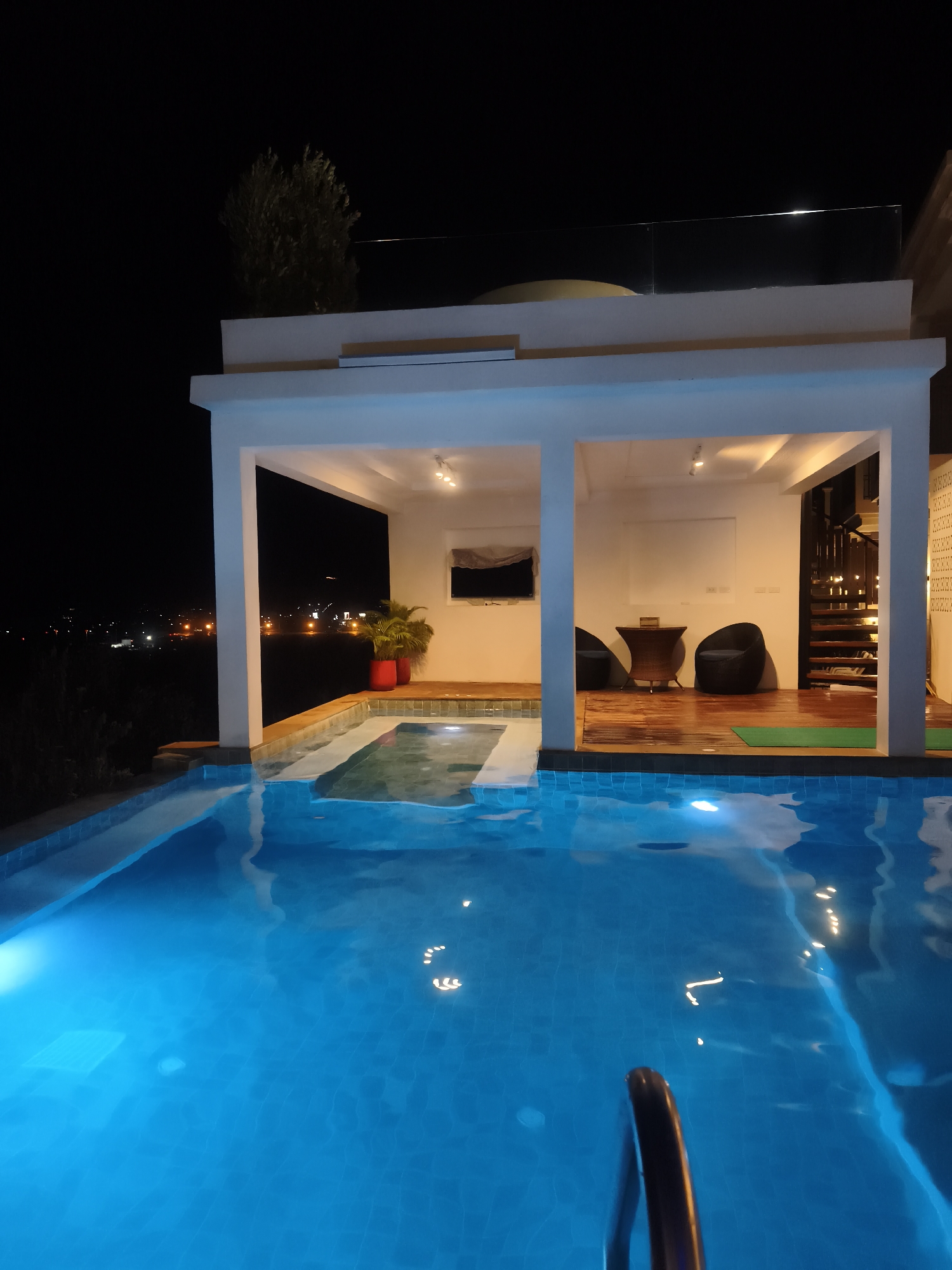 The image shows a swimming pool in บ้านบางรักษ์, Thailand, at night. The pool is brightly illuminated with blue light. Adjacent to the pool is a covered patio area with a wooden deck, containing seating, a small table, and a staircase. A potted plant is visible at the edge of the patio. In the background, city lights can be seen.