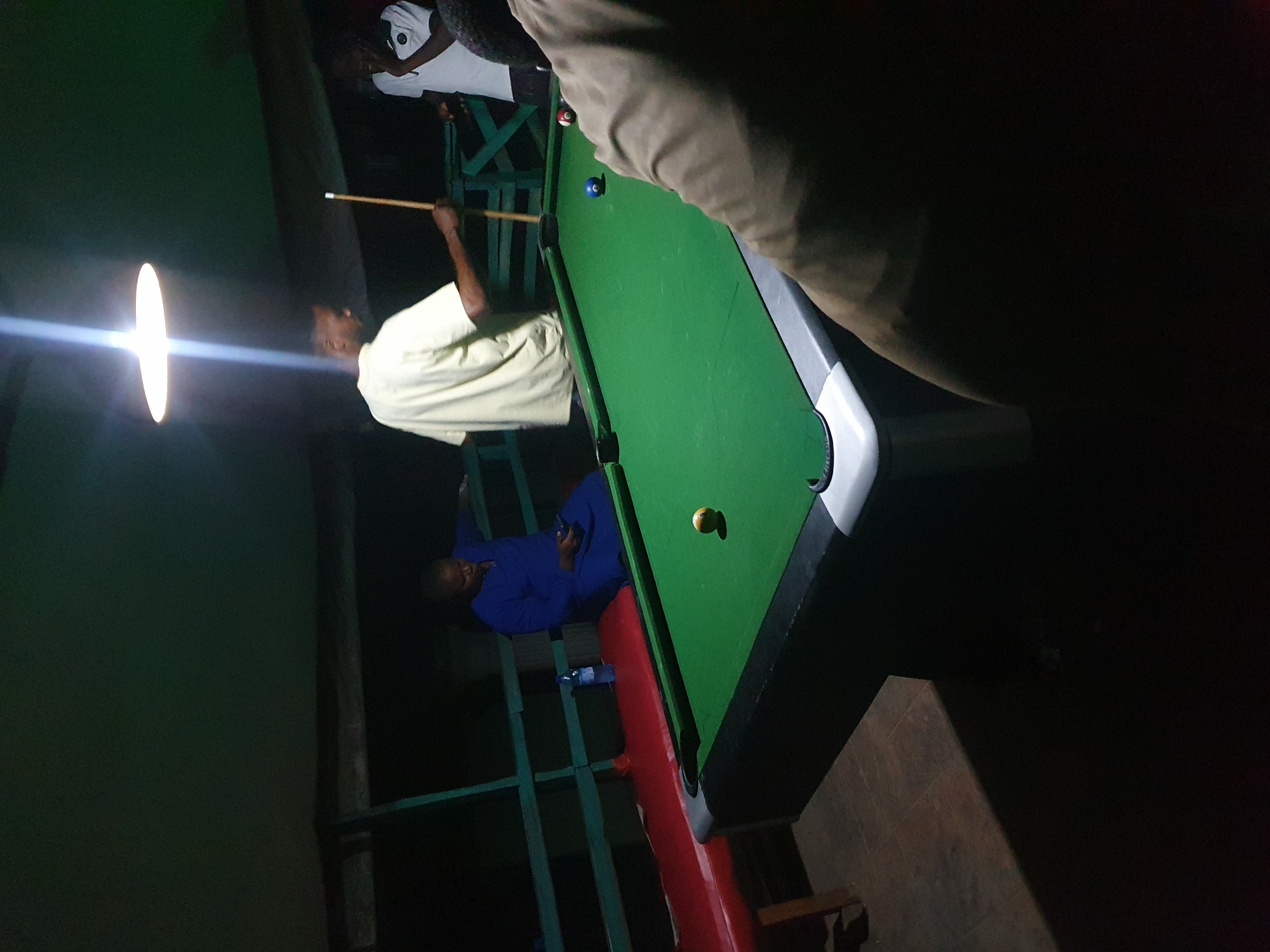The image depicts an outdoor nighttime setting, likely a social gathering or recreational area, potentially located in Lokoja, Nigeria, given the context. A pool table with green felt dominates the foreground, illuminated by a bright overhead light. There are several people present, though their features are not entirely clear due to the low light conditions.
In the center-left of the frame, a person in a blue garment sits on a red bench next to the pool table, holding a smartphone. To their right, a person dressed in a light-colored shirt stands by the pool table, holding a cue stick, appearing to be engaged in or preparing for a game of pool. Another individual in a white t-shirt is visible in the background on the right, partially obscured. The environment appears to be covered, possibly by a canopy or roof, with visible wooden structures and railings in the background. The overall atmosphere suggests a casual and communal gathering, possibly at a local bar, club, or private residence.