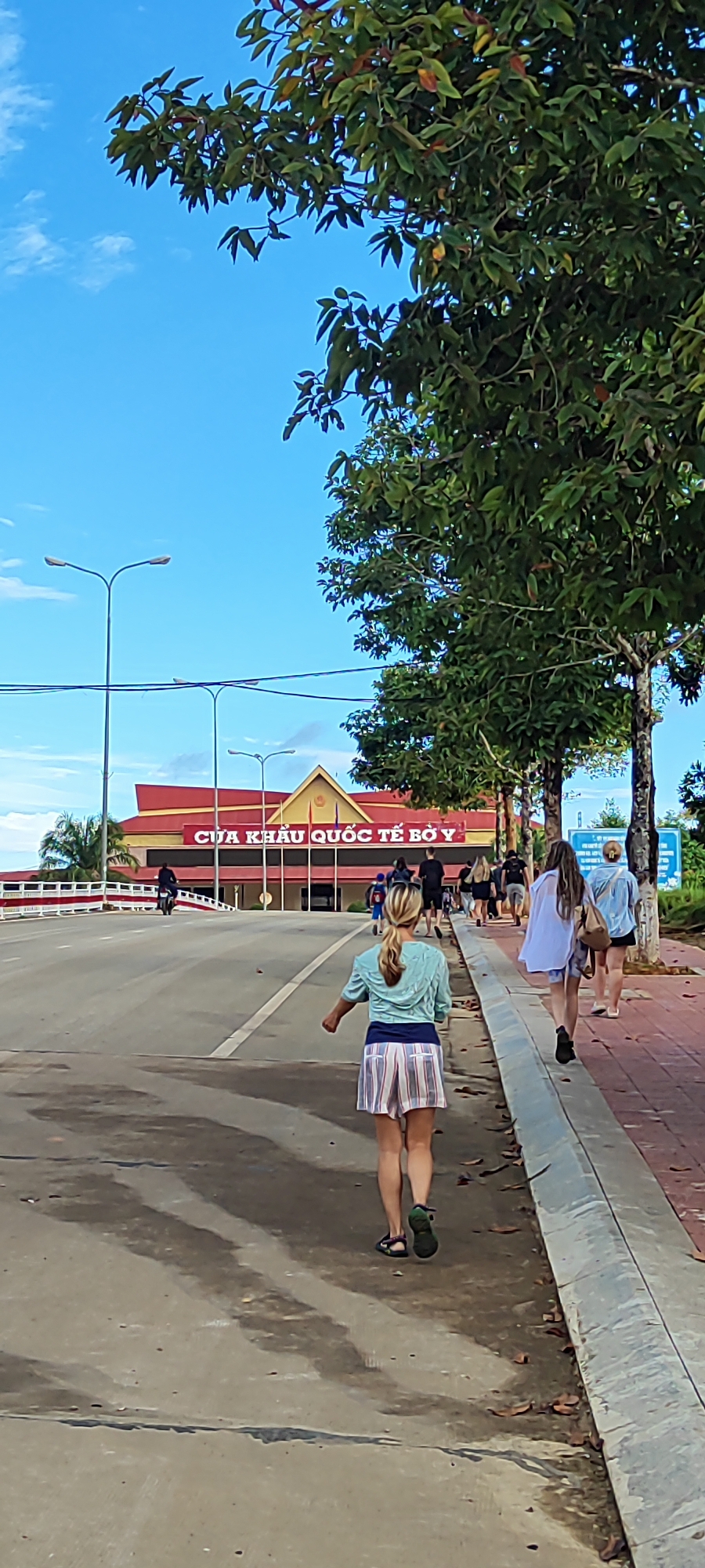 The image shows a street scene in Bờ Y, Vietnam, under a bright blue sky with scattered clouds. In the foreground, a woman with blonde hair tied in a ponytail is walking away from the camera on a paved road. She is wearing a light blue hooded jacket and a striped skirt. To her right, a sidewalk with a brick pattern is visible, lined with trees. Several people are walking on the sidewalk, heading towards a large building in the background.
The building has a red roof and a yellow facade with red lettering that reads "CỬA KHẨU QUỐC TẾ BỜ Y," indicating it is the Bờ Y International Border Gate. Streetlights are visible along the road. On the left side of the road, there is a white railing and more trees. A motorcycle is seen in the distance on the road, moving away from the border gate. The overall scene depicts a bustling border crossing with pedestrians and vehicles.