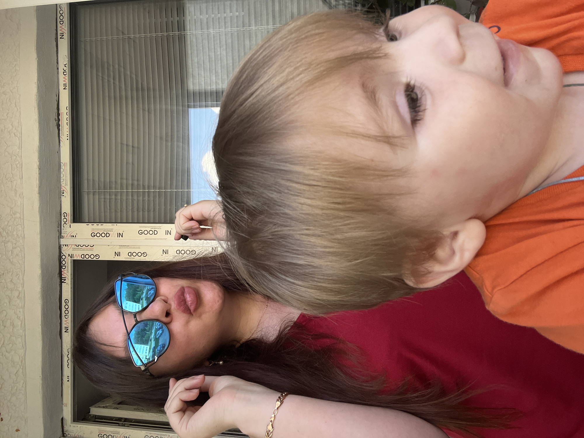 The image shows two people, a woman and a young child, appearing to be outdoors, possibly on a balcony or near a window. The woman is wearing sunglasses with blue mirrored lenses that reflect the surrounding environment, showing trees and vehicles. She has long, dark hair and is wearing a red top. The child has light-colored hair and is wearing an orange shirt. The child's face is close to the camera, with one eye visible. The background features a window with horizontal blinds. The scene suggests a casual, possibly daytime setting. The woman's expression, as partially visible, appears relaxed, and the overall mood seems light and familial. Some visible text on the window frame reads "GOOD WIN". The orientation of the image is landscape.