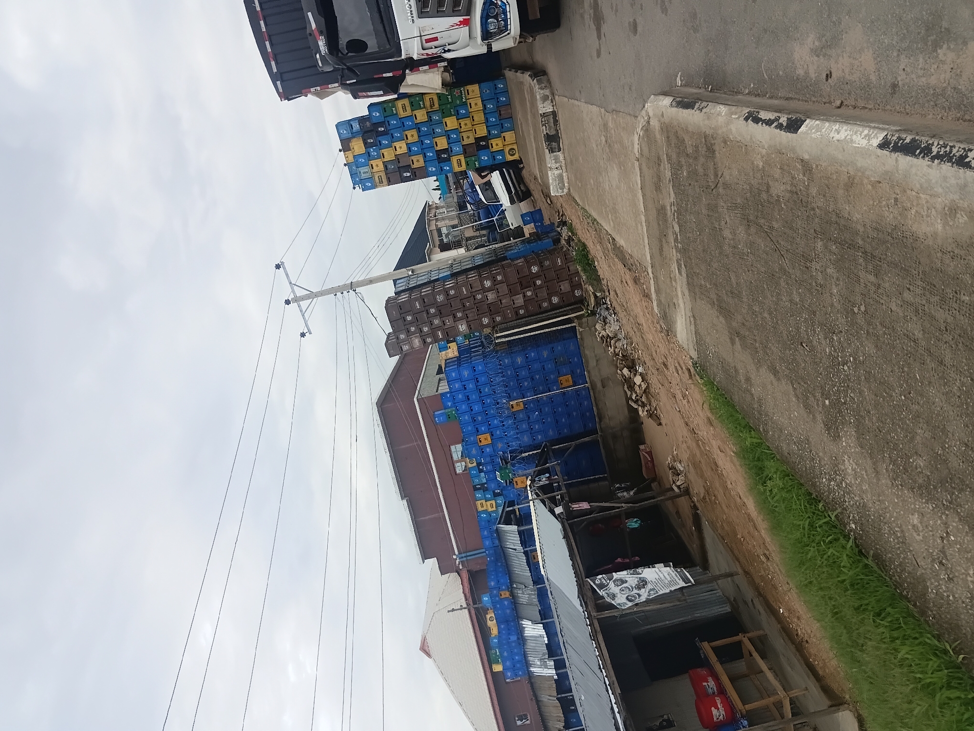 The image depicts a street scene in Jalingo, Nigeria, likely an outdoor market or distribution point. Towering stacks of blue and brown plastic crates are prominently featured, some filled with beverages, suggesting a commercial enterprise. A white truck is parked on the right, and the back of a dark truck is visible to its left. A small, open-front structure with a tin roof is situated on the left side of the image, with some red containers on a wooden table. In the background, a multi-story building with a brown facade and a lighter-colored roof is visible. The ground is a mix of dirt, gravel, and a paved concrete walkway. Overhead, power lines traverse the sky, which is overcast. The scene appears to be a bustling commercial area with goods being stored and possibly loaded or unloaded.