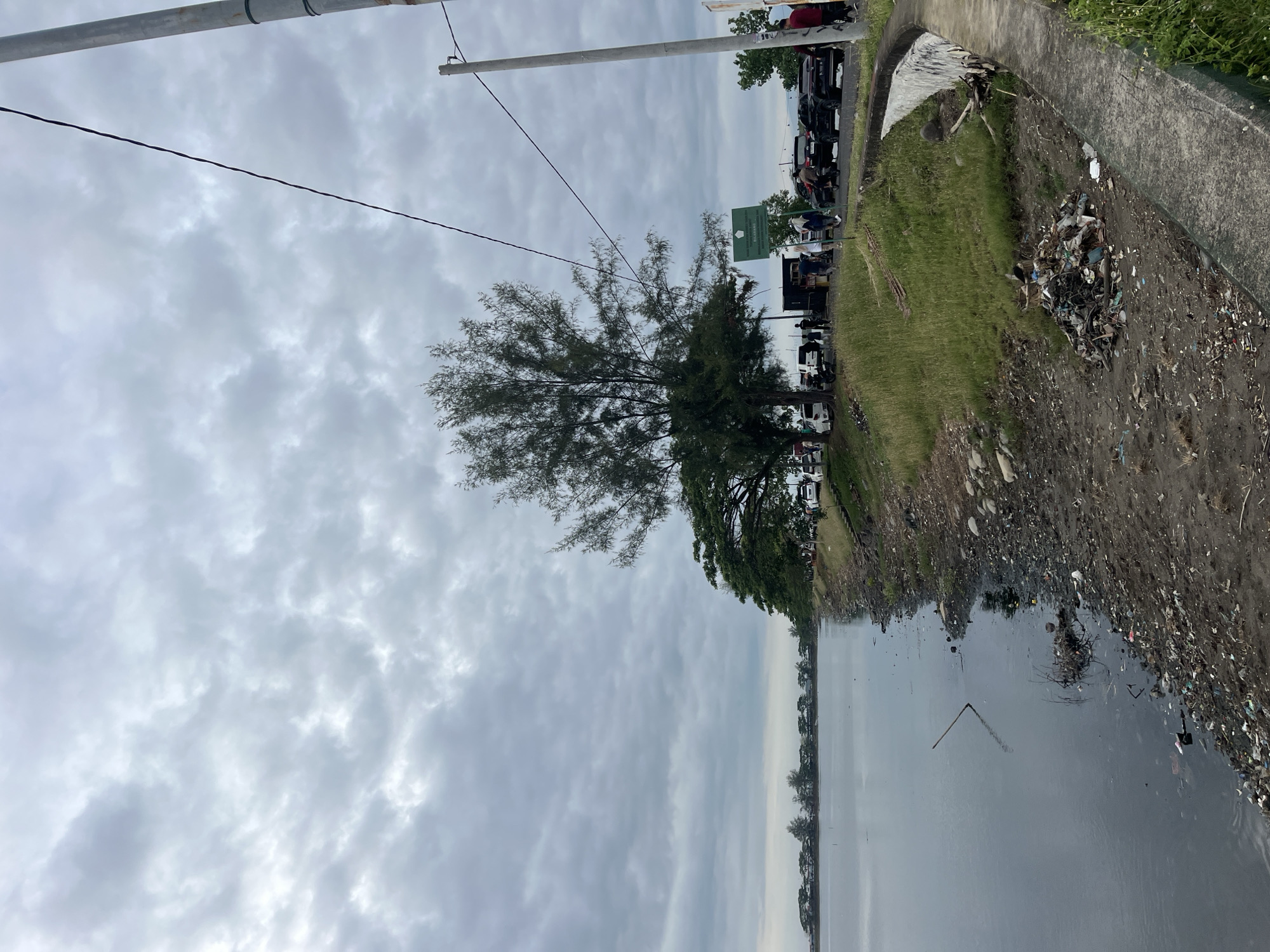 The image shows a polluted waterfront scene in Lhokseumawe, Indonesia. Overcast sky above, with an overhead power line extending across the upper portion of the image. A tree stands prominently in the center, partially obscuring parked vehicles including cars and motorbikes. A green sign is present near the vehicles. To the right, a concrete walkway lines the waterfront. Debris and trash are heavily accumulated along the embankment, extending into the water, which is dark and murky. The scene lacks distinct people or actions, emphasizing environmental pollution.
