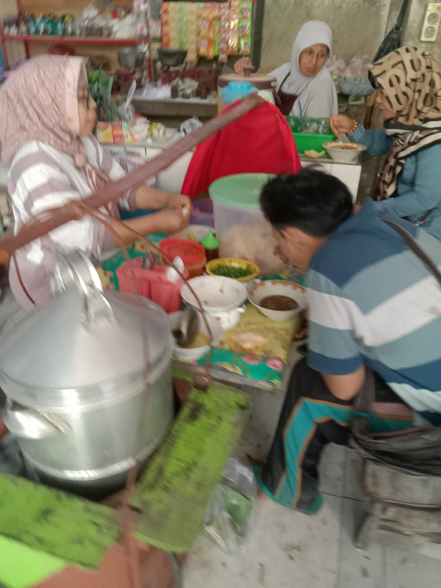 In Sugihwaras, Indonesia, a busy market scene unfolds. Three individuals are visible. To the left, a woman wearing a pink headscarf and a striped shirt is partially obscured by a large, silver steaming pot. She is holding a long, wooden utensil. In the center background, a woman in a white headscarf and apron is preparing food. To the right, a woman with a patterned headscarf and a blue top is seated, stirring a bowl of food. The table is laden with various bowls, containers, and what appears to be food items. The environment suggests an outdoor food stall or market setting.