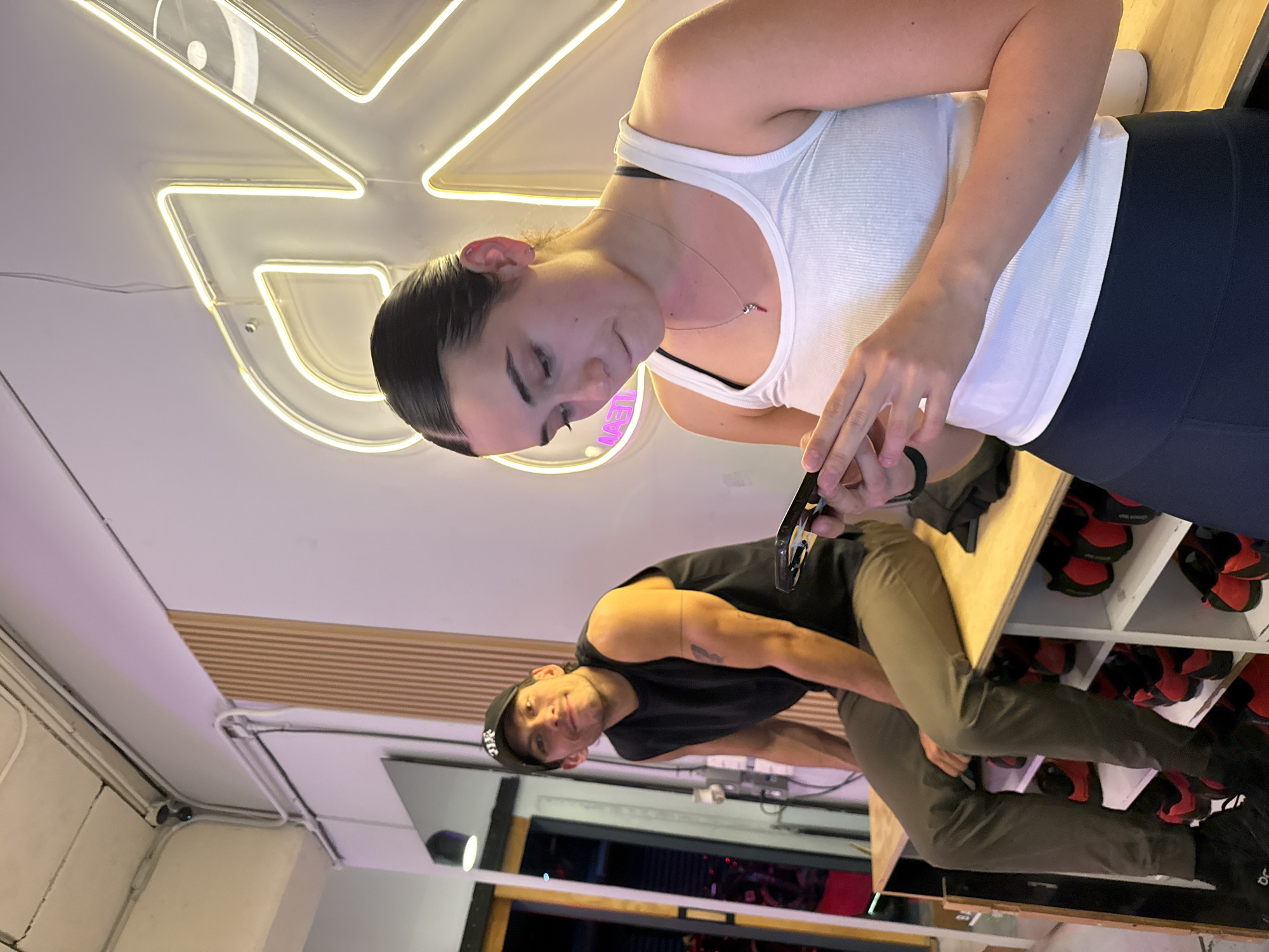 Two individuals are present in an indoor environment. A fair-skinned female with dark hair pulled back is positioned in the upper portion of the image, wearing a white tank top and dark bottoms. She is holding a black object. In the lower portion of the image, there is a fair-skinned male wearing a black tank top, green pants, and a black baseball cap. He has tattoos on his arm. They appear to be standing near a wooden surface that is possibly a shelf containing red and black shoes. The background features a ceiling with exposed electrical conduit and a neon light fixture. The lower part of the background contains a glass window looking out on an interior with a red and black design. No specific details suggest this is in Puebla, Mexico.