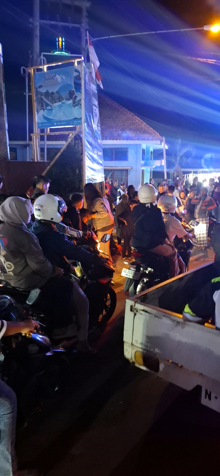 This is a medium-shot, vertical, night-time photograph of a crowd gathered on a street in Prigen, Indonesia. In the foreground, several individuals are seated on motorcycles. One motorcycle, in the lower left, has a rider wearing a grey hooded jacket. To the right of this, another motorcycle has a rider wearing a white helmet and dark jacket. Behind these, a white pickup truck is visible, with its rear section occupied by at least one person wearing yellow sneakers.
In the mid-ground, a larger group of people are standing and interacting. Several individuals are wearing helmets, indicating they are likely on motorcycles. One person in the center of the frame, wearing a light-colored jacket, appears to be interacting with someone on a motorcycle. Motorcycles are parked throughout the scene.
In the background, a building with a tiled roof is illuminated by blue and white lights. A banner with an image of mountains and water is displayed vertically on a frame, with an Indonesian flag attached. Above this, a structure topped with green and orange lights is visible, possibly a minaret. The overall environment suggests a lively gathering or event taking place on a street at night.