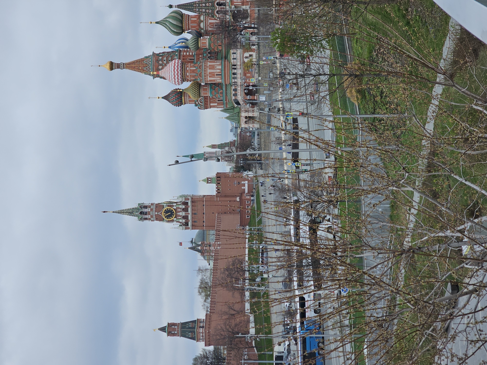 The image captures a cityscape in Moscow, Russia. It prominently features the Moscow Kremlin with its brick walls and various towers topped with green roofs and spires. The Spasskaya Tower, recognizable by its large clock face, is visible. Adjacent to the Kremlin stands the colorful St. Basil's Cathedral, known for its iconic onion-shaped domes. A street with traffic runs between the viewer's perspective and the buildings, bordered by green spaces and pedestrian walkways. Bare tree branches partially obscure the view, suggesting it may be early spring. Vehicles, including a blue bus, are seen on the road. Pedestrians are also visible along the street and walkways.