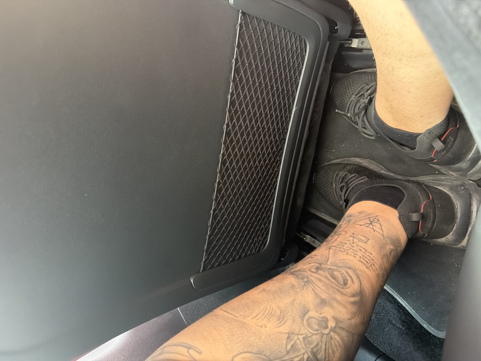 The image shows a close-up, high-angle view of a person's legs and feet inside a car in Trastenik, Bulgaria. The setting appears to be daylight.
The central subject is a person's legs and feet. The leg on the left side of the frame (viewer's left) is extensively covered with black and grey tattoos. These tattoos include what appears to be a large, intricate face or lion's head, an area of small, illegible text or script, and geometric patterns, possibly including a pyramid-like structure. This leg is smooth. The person is wearing a black athletic sneaker with black ankle socks.
To the right, part of the person's other leg is visible, which appears hairy and is also wearing a similar black sneaker and black socks. Both feet rest on a dark, possibly black, car floor mat that shows some minor dust or debris. In the background, sections of car seat rails are visible.
The car's interior is predominantly dark. On the left side of the frame, a black, textured car door panel is visible, featuring a black mesh storage pocket or net. The overall impression is of a casual scene within a vehicle. No specific activity beyond sitting is evident, and no emotions can be discerned. The tattooed script is not clearly legible.
