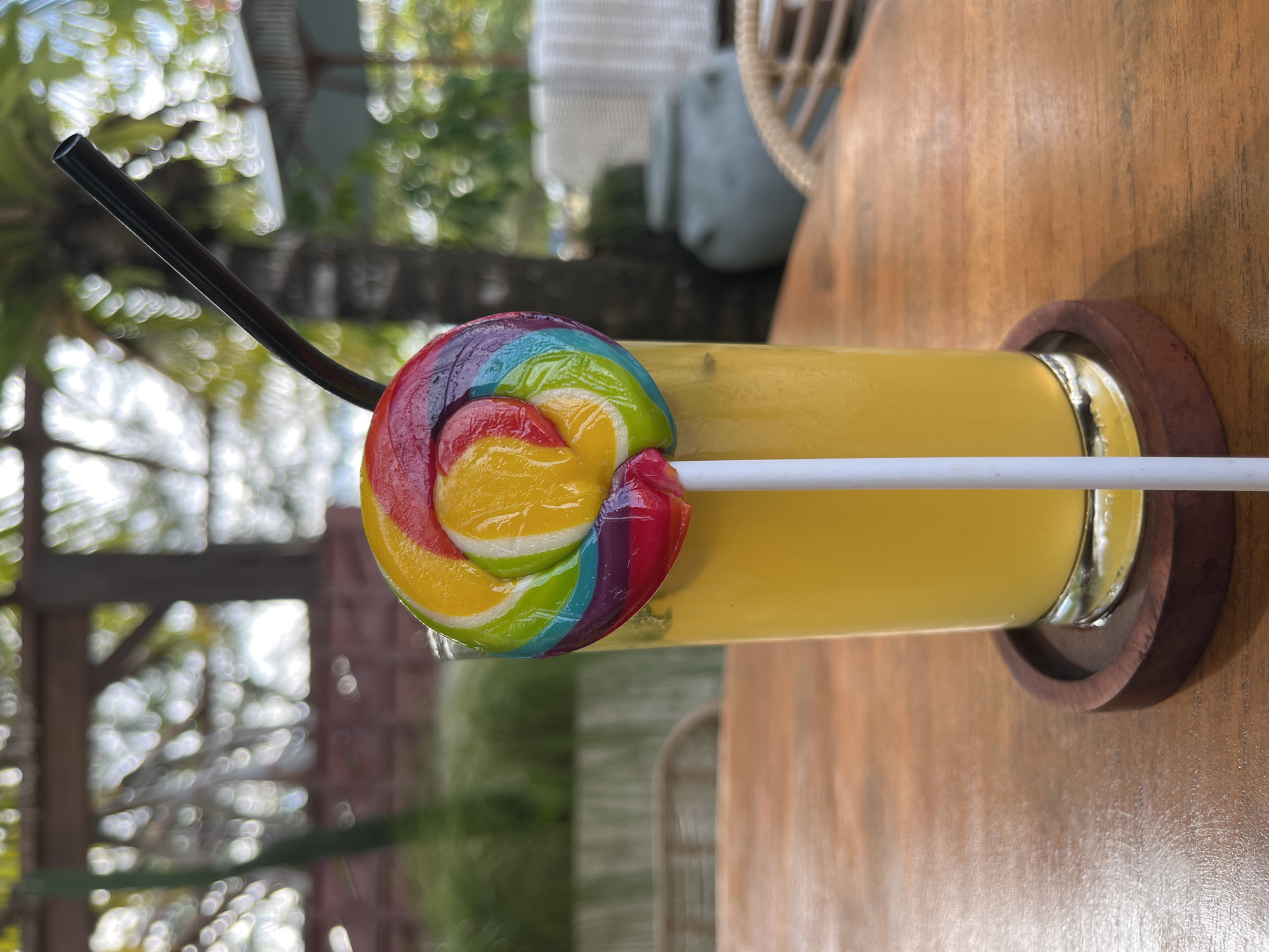 A rainbow swirl lollipop is stuck into a tall glass of yellow liquid, possibly juice or a cocktail, which sits on a wooden table. A black straw is emerging from the glass. The background is blurred, showing greenery, palm leaves, and possibly outdoor seating with wicker furniture. The overall impression is a tropical setting, potentially a cafe or restaurant in Kuta Selatan, Indonesia, given the foliage and lighting. The image is taken from a slightly low angle, focusing on the drink and lollipop.