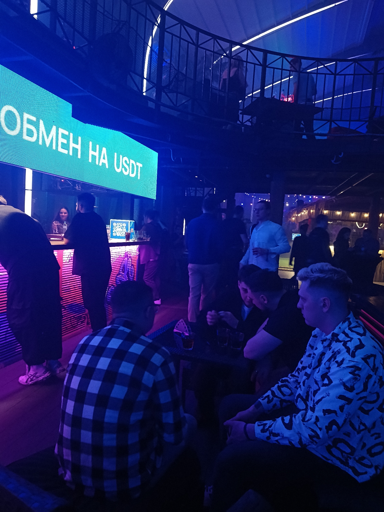The image shows a bar or club in Moscow, Russia. The sign above the bar reads "Обмен на USDT" (Exchange for USDT). People are at the bar and seated at tables. The environment is dimly lit with blue and purple lighting. There are people on an upper level balcony. There is a QR code on the bar.