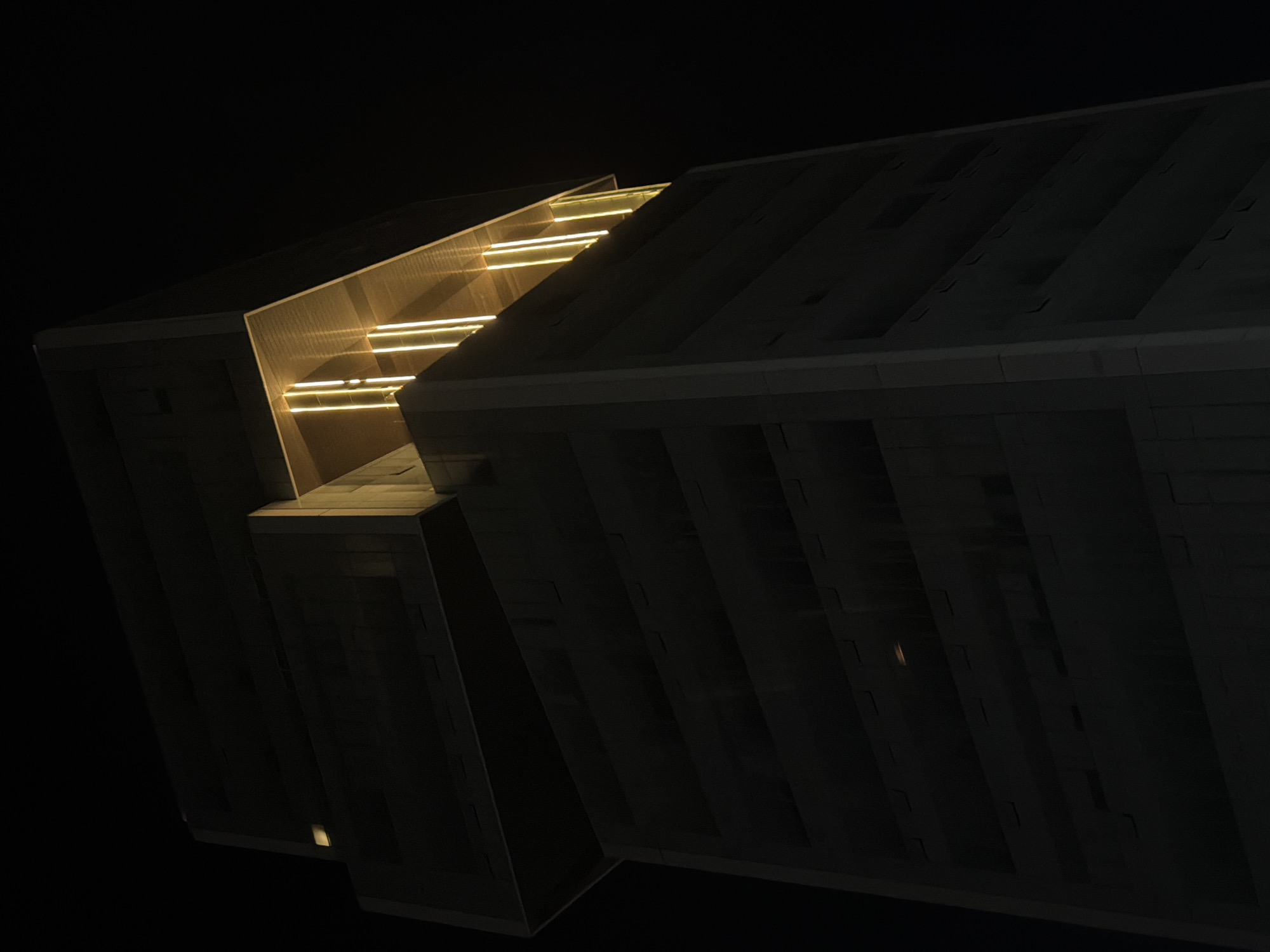 The image displays a modern architectural structure at night, set against a completely dark background. No people are visible in the scene. The building, likely located in Puebla, Mexico, is characterized by its geometric, angular design and imposing presence.
A significant portion of the structure is shrouded in deep shadow, enhancing its dramatic silhouette. However, a prominent section on the left side of the building is brightly illuminated by several parallel, horizontal strips of warm yellow-orange light. These lights are integrated into the architecture, highlighting an inclined, possibly stepped or terraced, inner surface that appears to be made of a textured, light-colored material. The interplay of light and shadow emphasizes the building's sharp lines and volume.
The darker sections of the building reveal subtle details of its exterior, including what appear to be vertical divisions, panels, or recessed features, suggesting a complex facade. The overall aesthetic is one of contemporary design, evoking a sense of grandeur and mystery. The scene focuses solely on the architectural form and its dramatic nighttime illumination. There is no visible text in the image.
