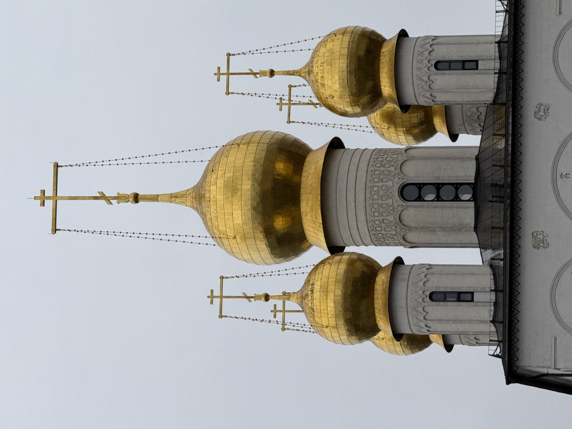 The image shows a row of golden domes atop the Cathedral of the Archangel, located within the Kremlin in Moscow, Russia. The domes are topped with crosses, and there are chains strung between them. The architecture is predominantly white stone with detailed patterns around the base of the domes. The sky is overcast and grey.