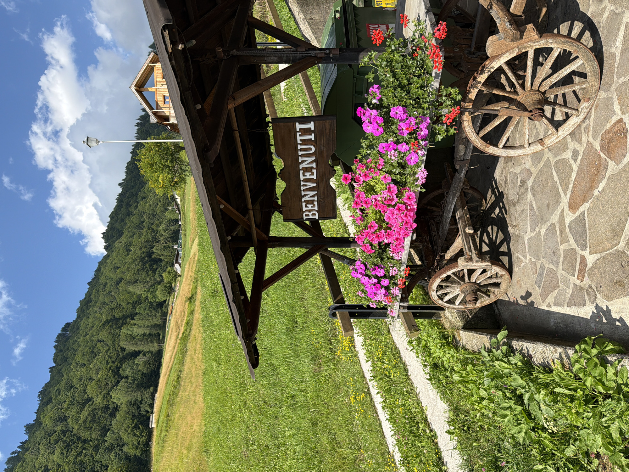 The image depicts a rustic wooden wagon filled with vibrant pink and red flowers, serving as a decorative element. A sign hanging from a wooden structure above the wagon reads "BENVENUTI," which means "welcome" in Italian. The wagon is situated on a paved stone area. In the background, a lush green hillside slopes upwards, covered with trees and patches of golden-yellow grass. A modern wooden building with a triangular roof and a street lamp are visible on the hillside. The sky above is blue with scattered white clouds, indicating a clear day. The scene appears to be an outdoor setting, possibly a welcoming area for a rural establishment in the mountainous region of Sauris / Zahre, Italy, given the landscape and the Italian signage.