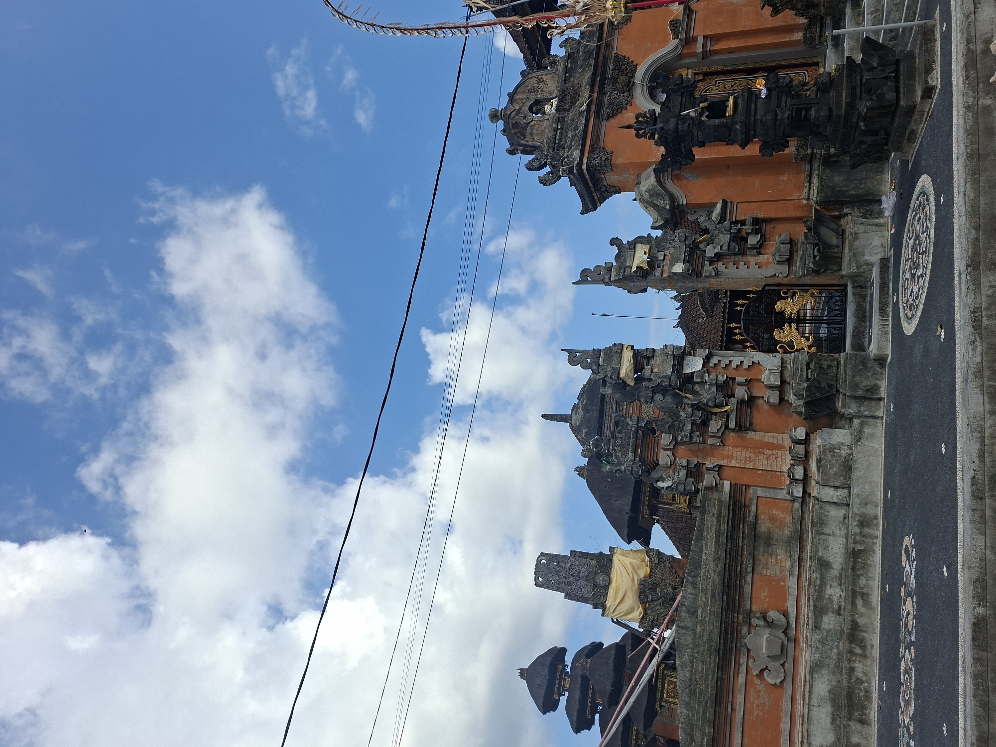 The image shows the ornate entrance to a temple in Blahbatuh, Indonesia. The temple features traditional Balinese architecture with intricate stone carvings, orange brick walls, and thatched roofs. A golden gate with lion motifs stands prominently in the center. The foreground consists of a paved pathway with decorative mosaic patterns. The sky above is blue with scattered white clouds, and power lines are visible. The scene appears to be a public space, possibly a tourist attraction.
