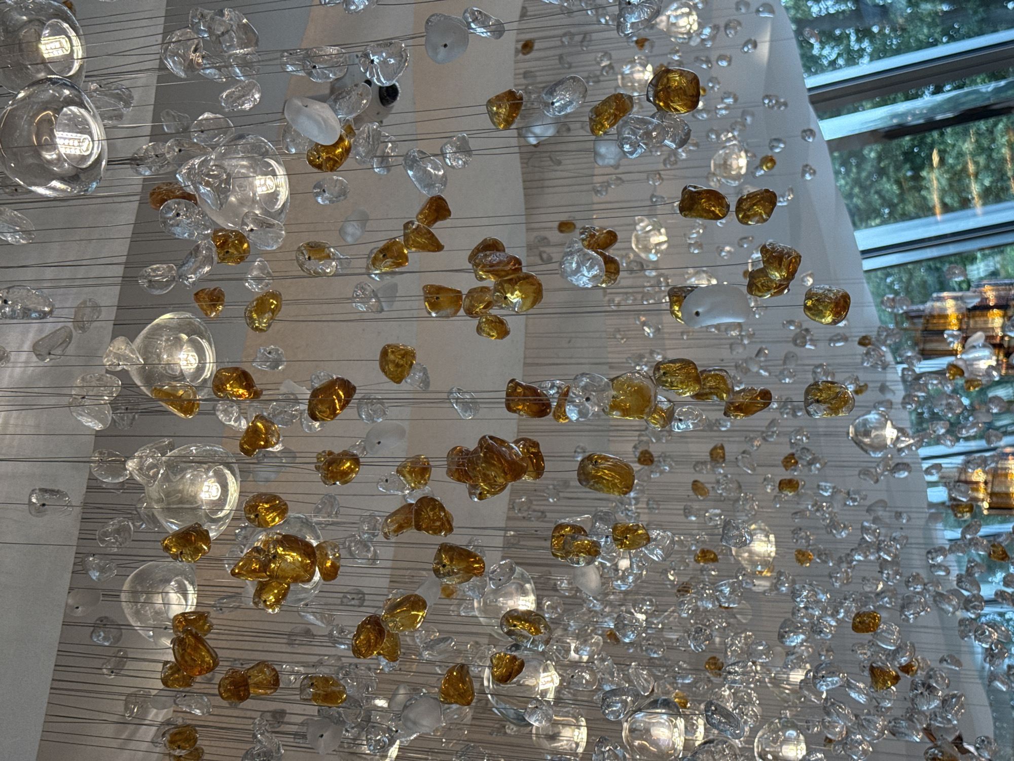 The image shows a close-up view of a large, modern chandelier made of many suspended glass elements. The elements vary in size and shape, with some appearing clear and others having a warm, amber hue. Many of the clear elements are shaped like irregular pebbles or teardrops, while some of the amber elements resemble polished stones. Interspersed among these are clear glass spheres that appear to contain light bulbs, suggesting they are functional light fixtures. The chandelier is hanging from a white ceiling, and the elements are arranged in a dense, cascading pattern. The background is out of focus, but suggests a bright, natural light source and greenery, possibly trees seen through a window. No people are visible in the image. The style of the chandelier suggests a contemporary interior design, common in upscale hotels, restaurants, or public spaces. The request to include the city of Nizhny Novgorod, Russia is not supported by any visual information within the image.