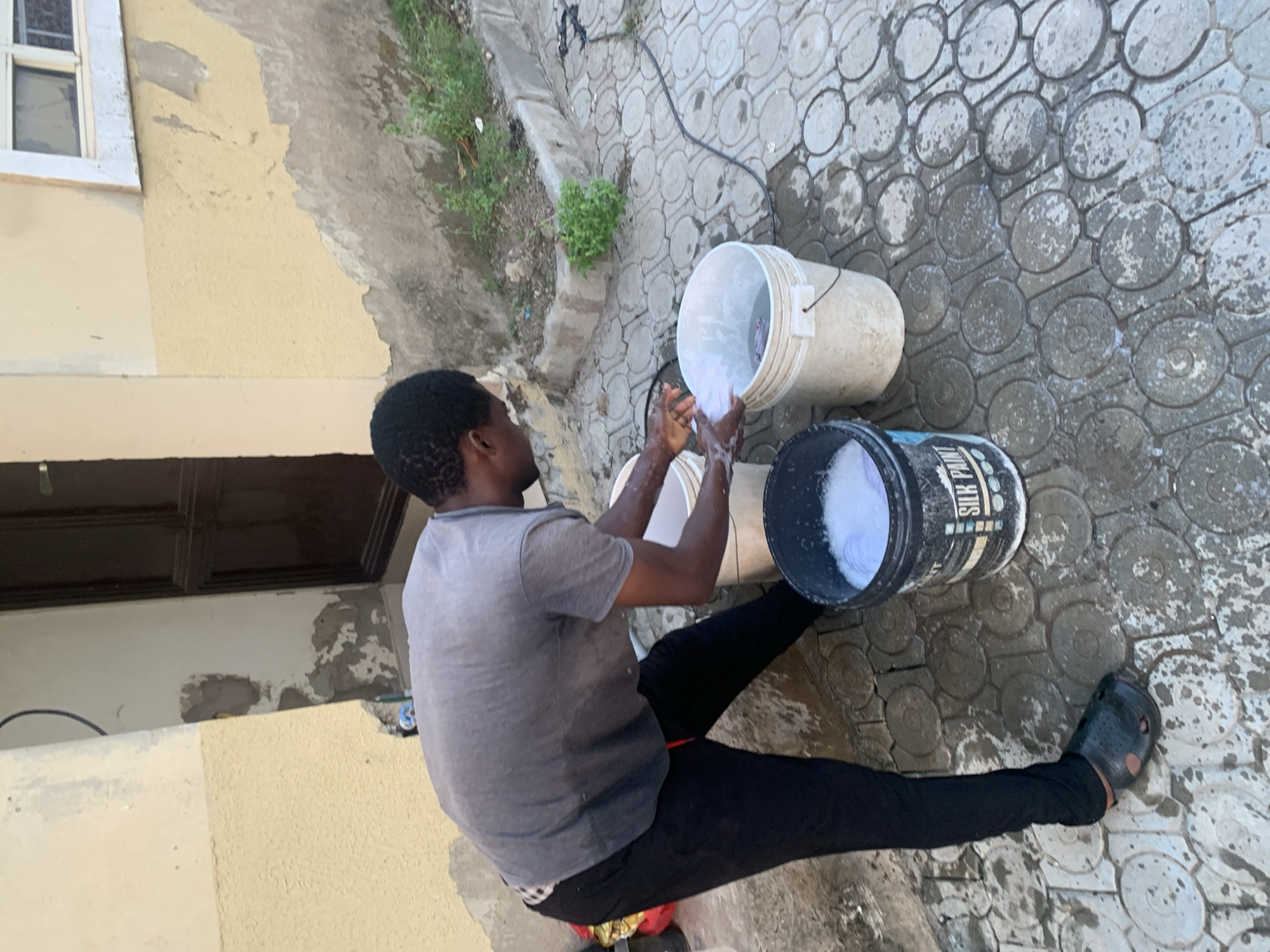 A person is washing clothes outdoors. They are wearing a grey t-shirt and black trousers. The person is sitting on a red object on the ground, with their legs crossed. In front of them are two buckets. One is white and appears to contain water and some white fabric. The person is holding their hands in this bucket, suggesting they are washing. The second bucket is black and has some white foamy substance inside, possibly detergent or soap.
The setting appears to be a paved area with circular patterned concrete tiles. There is some greenery to the left of the scene, and a building with a yellow-painted wall and a window is visible in the background. The ground is wet, indicating recent rain or the ongoing activity of washing.
The scene suggests a typical outdoor chore, possibly taking place during the daytime. There are no strong emotional cues visible, but the person appears focused on the task. There is text visible on the black bucket that reads "SILK PAINTS". The overall impression is of a practical, everyday activity in an outdoor environment in Abuja, Nigeria.