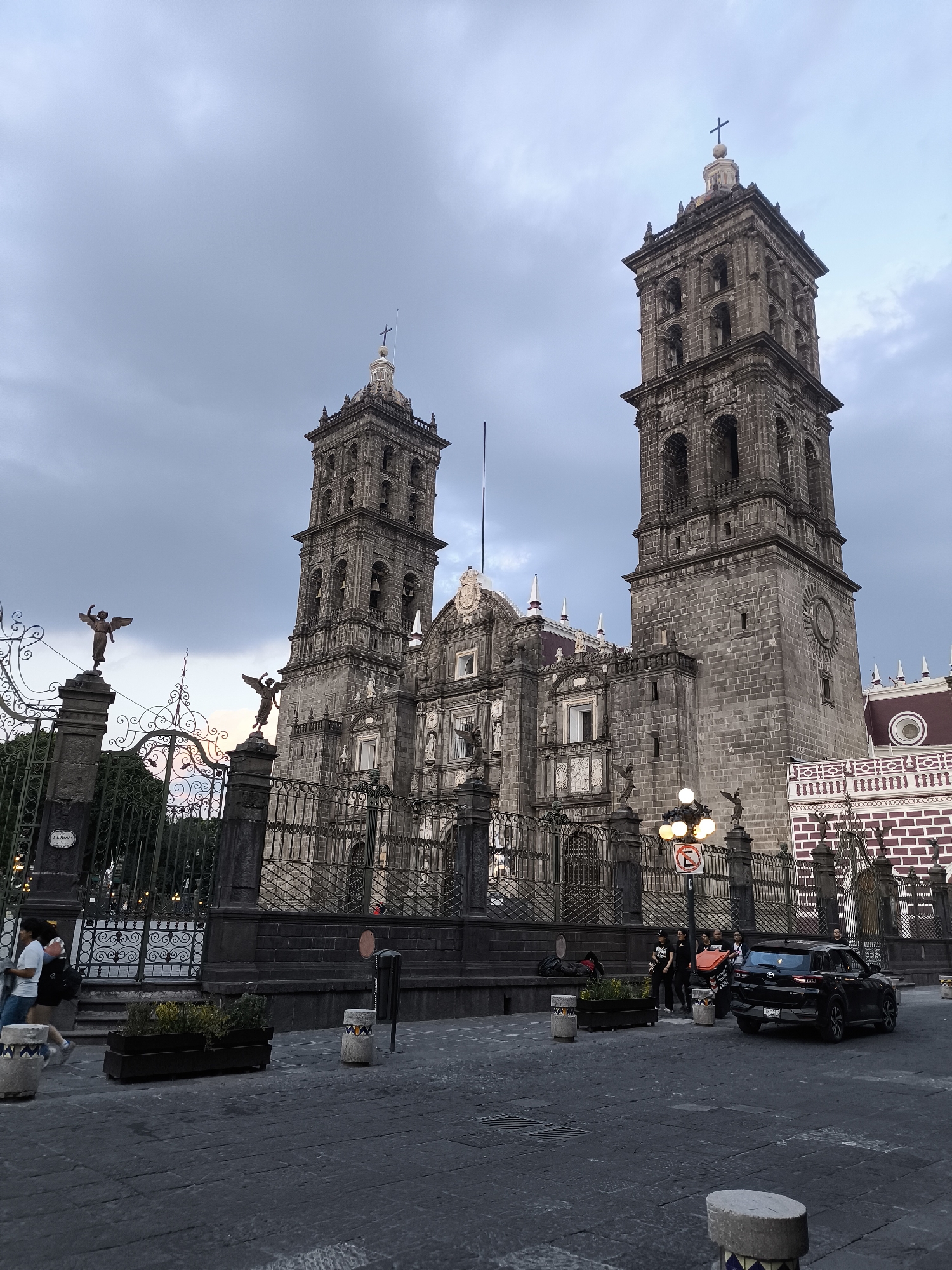 The image shows the Puebla Cathedral, a major religious structure located in the city of Puebla, Mexico. The cathedral is constructed of dark gray stone and features two tall bell towers topped with crosses. The building is enclosed by an ornate wrought iron fence, decorated with statues of angels.
In the foreground, there's a paved public square with several bollards and planters. To the right, a black SUV is parked, with a group of people nearby. Another group of people is gathered near the fence. A couple is walking in the left foreground.
The sky is cloudy, suggesting either early morning or late afternoon.