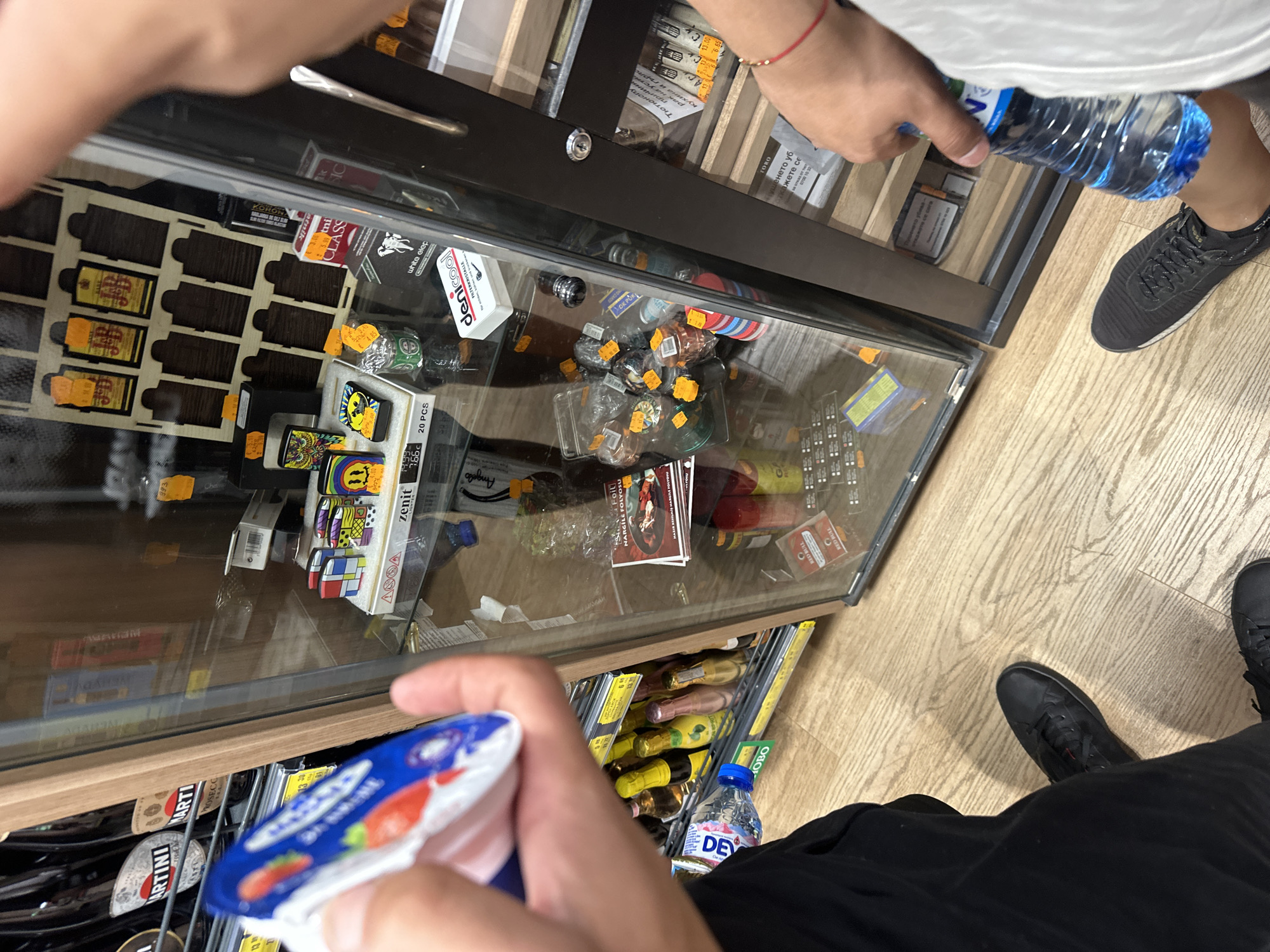 This is a photograph taken inside a convenience store in Ruse, Bulgaria. The perspective is from a person holding a yogurt cup and a bottle of water. The store features a glass-fronted display cabinet filled with various items, including lighters, small bottles of alcohol, and packaged goods. The floor is made of light-colored wood planks. On the left side of the frame, a shelf displays rows of small bottles, some of which appear to be alcoholic beverages like champagne or wine. The person's legs and feet are visible at the bottom of the image, wearing black trousers and black sneakers. The display cabinet's shelves hold a variety of products, some with price tags. The overall lighting suggests an indoor retail environment.