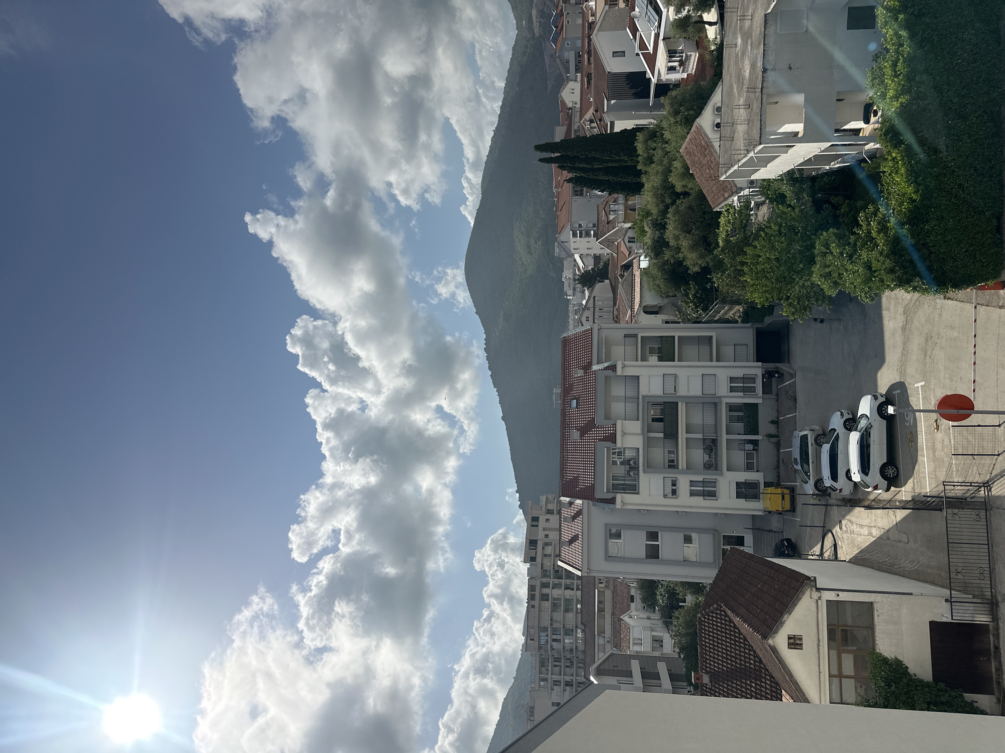 This is an outdoor daytime shot looking down from a higher elevation. The scene includes the sky, a mountain, and urban structures.
The sky is predominantly blue with bright sunlight at the bottom left, with puffy white clouds occupying much of the left side of the image. A verdant, steep mountain rises vertically in the center of the frame.
To the right of the mountain, there is a dense grouping of buildings, which appear to be residential apartments and houses. These structures are mostly white, with some having red-tiled roofs. Trees are interspersed among the buildings, adding greenery. A street-level parking area is visible with several parked vehicles including three white cars and one small yellow car. A stop sign is installed on the right edge of the road.
Based on the architectural style and landscape features, the location can be identified as Budva, Montenegro.