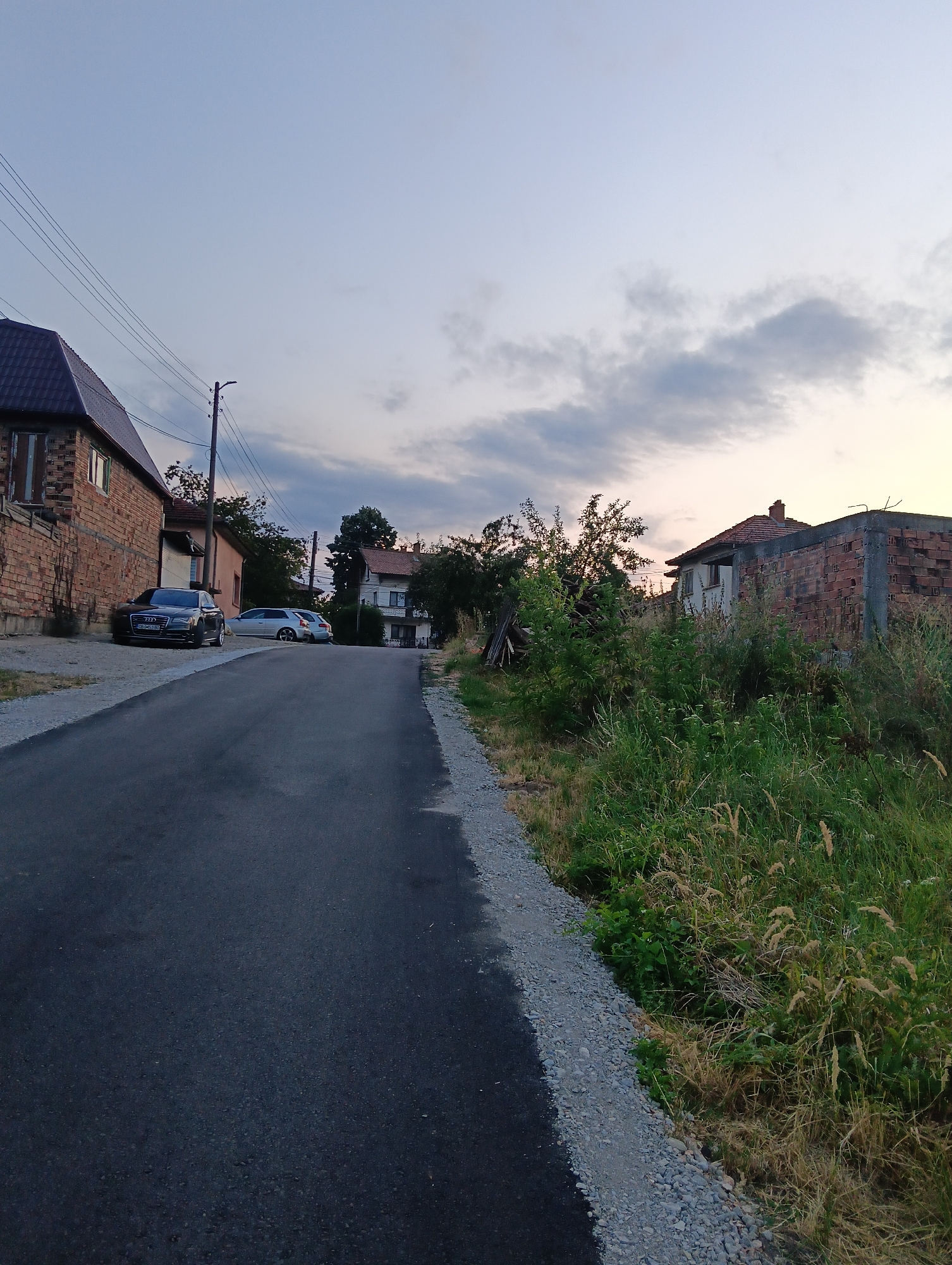 The image depicts a street scene in Oreshak, Bulgaria. A paved road occupies the foreground, curving to the right and bordered by gravel on the left. On the left side of the road, a brick house with a dark tiled roof is visible. Two cars are parked on the side of the road in front of the house; a black Audi sedan and a silver station wagon. Further down the street, on the right side, a partially constructed brick building stands next to a wall made of exposed brick. Overgrown grass and weeds are abundant on the right side of the image. In the background, several houses and trees are visible, set against a sky with scattered clouds. The overall scene suggests a rural or suburban setting during dusk.