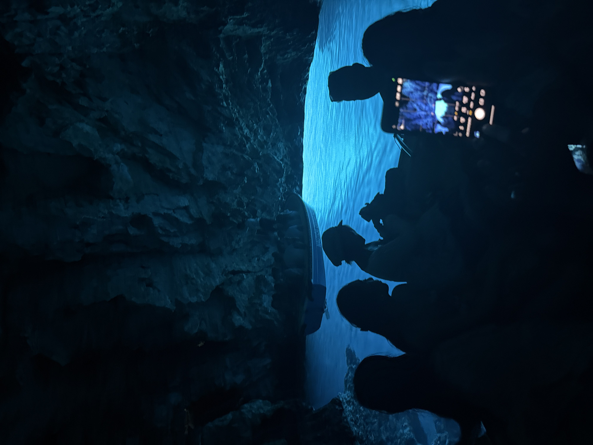 The image depicts the interior of the Blue Cave, a sea cave located off the coast of the island of Vis, near the town of Komiža, Croatia. The cave is illuminated by a surreal, vibrant blue light that reflects off the water, creating a magical atmosphere. Silhouetted figures of people are visible in the foreground, seated in a boat, their forms dark against the luminous blue water. One person in the foreground is holding up a smartphone, capturing the scene. The cave's rocky walls are visible in the background, dimly lit by the same ethereal blue glow. The overall impression is one of a captivating natural wonder, experienced by visitors in a boat.