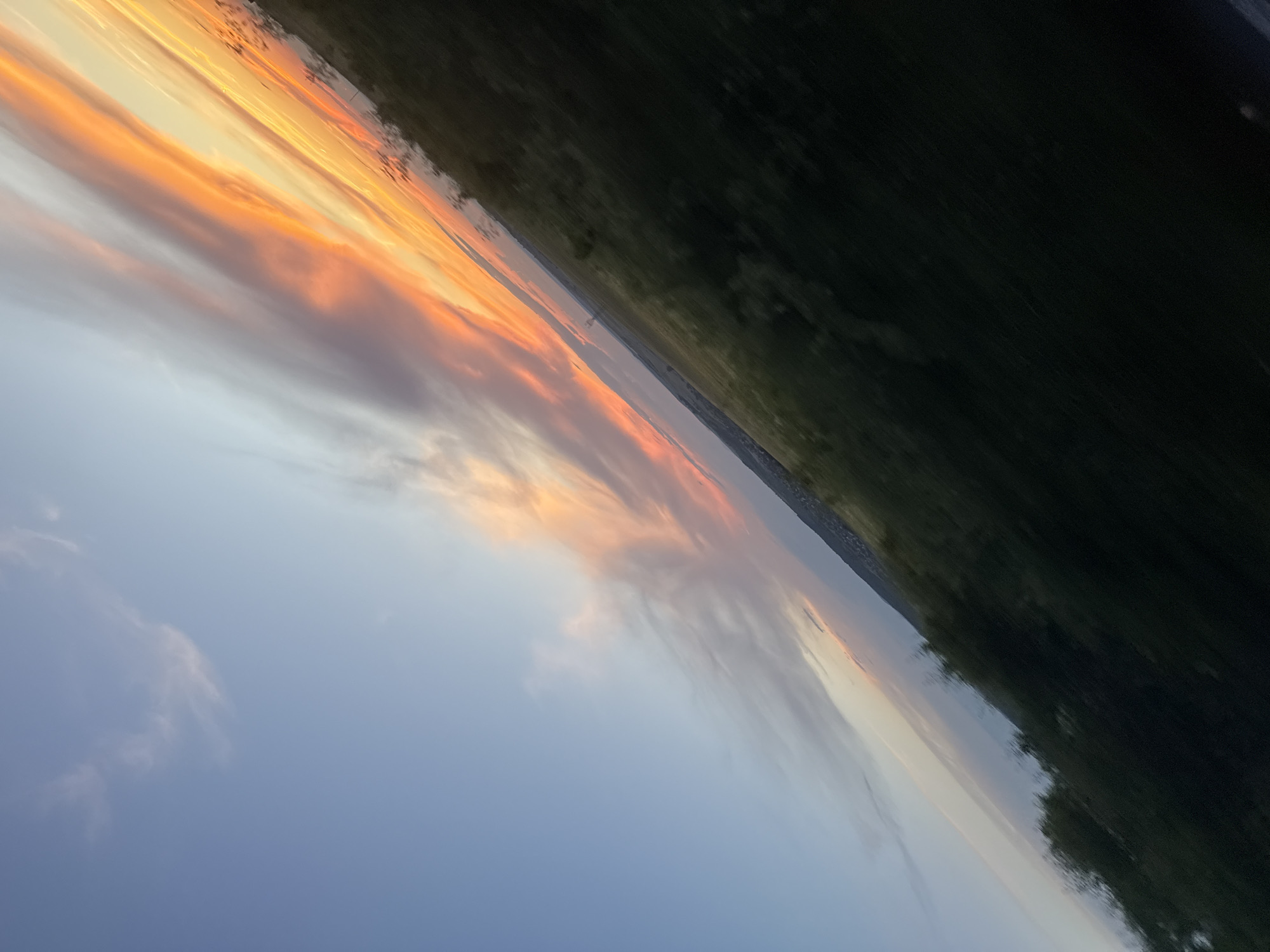 A photograph captures a diagonal view of a sunset over a landscape that could be near Izvor, Bulgaria. The sky is filled with dramatic clouds, painted in vibrant shades of orange, yellow, and pink, transitioning to a soft blue on the left side of the frame. Streaks of light and darker, wispy clouds create a dynamic visual. The lower portion of the image shows a dark, green landscape that appears to be fields or vegetation, with some blurry motion suggesting the photograph was taken from a moving vehicle. A faint line of trees or distant structures can be seen along the horizon.