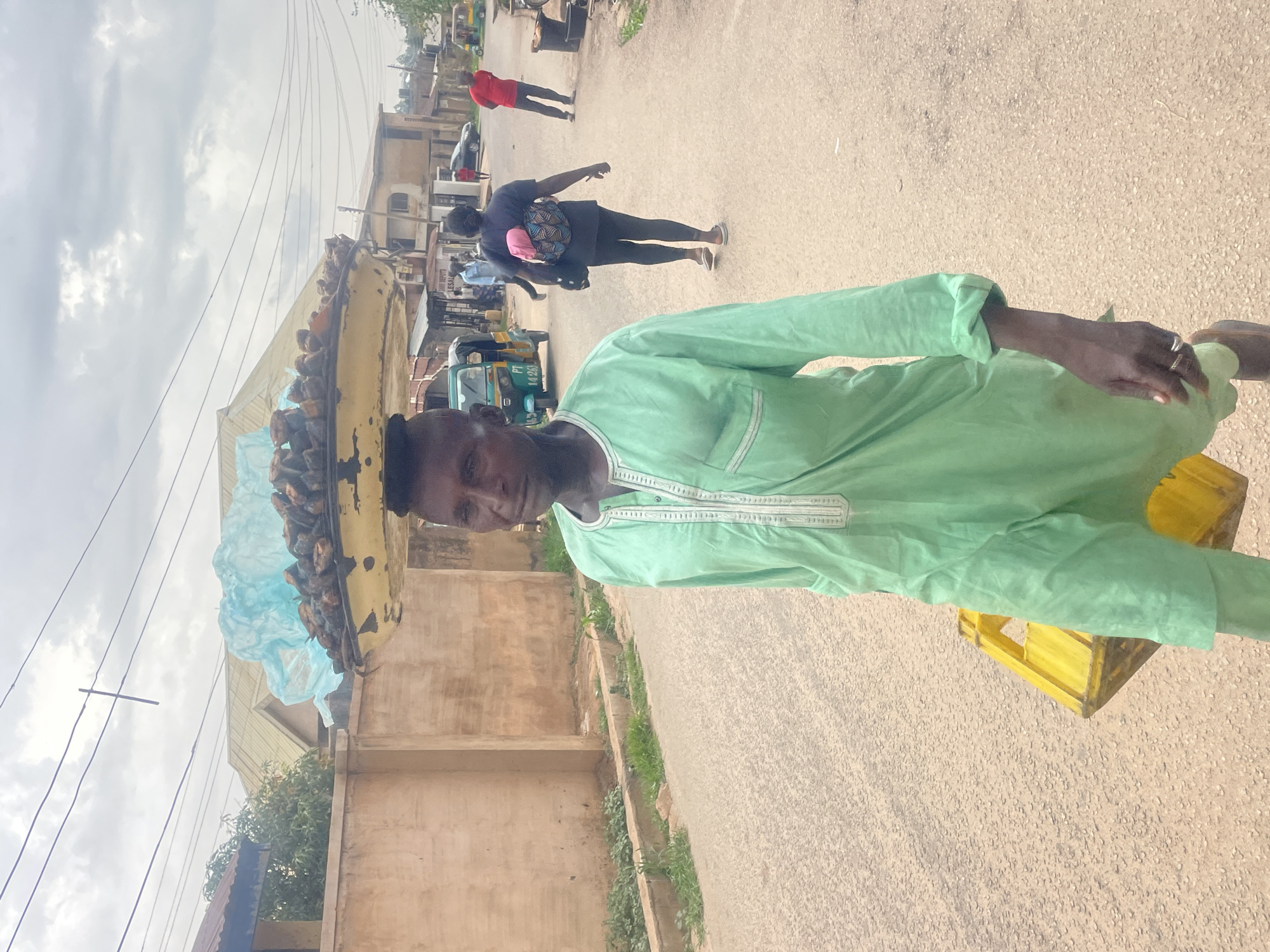 In Jos, Nigeria, a man wearing a light green, long-sleeved shirt walks along a paved road, carrying a large yellow tray filled with dried fish on his head, partially covered with a light blue plastic bag. He also carries a yellow crate in his right hand. Behind him, other pedestrians walk down the road. Buildings and vegetation line the side of the road. Overhead, power lines stretch across a cloudy sky.