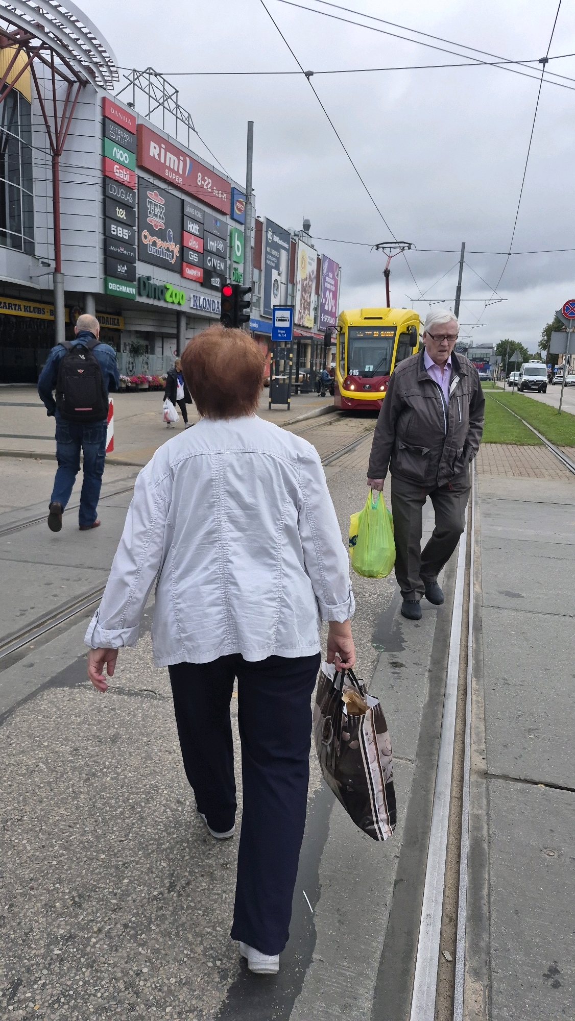 In Daugavpils, Latvia, on a cloudy day, three people are walking along a street with tram tracks. In the foreground, a woman with short red hair is walking away from the camera, wearing a light-colored jacket and dark pants, carrying a patterned shopping bag. To her right, a man in a brown jacket and gray pants is walking towards the viewer, holding a bright green shopping bag and looking to his right. Further in the background, a man with a backpack is walking away from the camera, wearing a denim jacket and blue jeans. A yellow tram is visible in the middle ground, stopped at a tram stop. The buildings behind the tram have various advertisements and signs, including "Rimi" and "Dino Zoo". The street is paved and shows signs of wear, with tram tracks embedded in the surface.