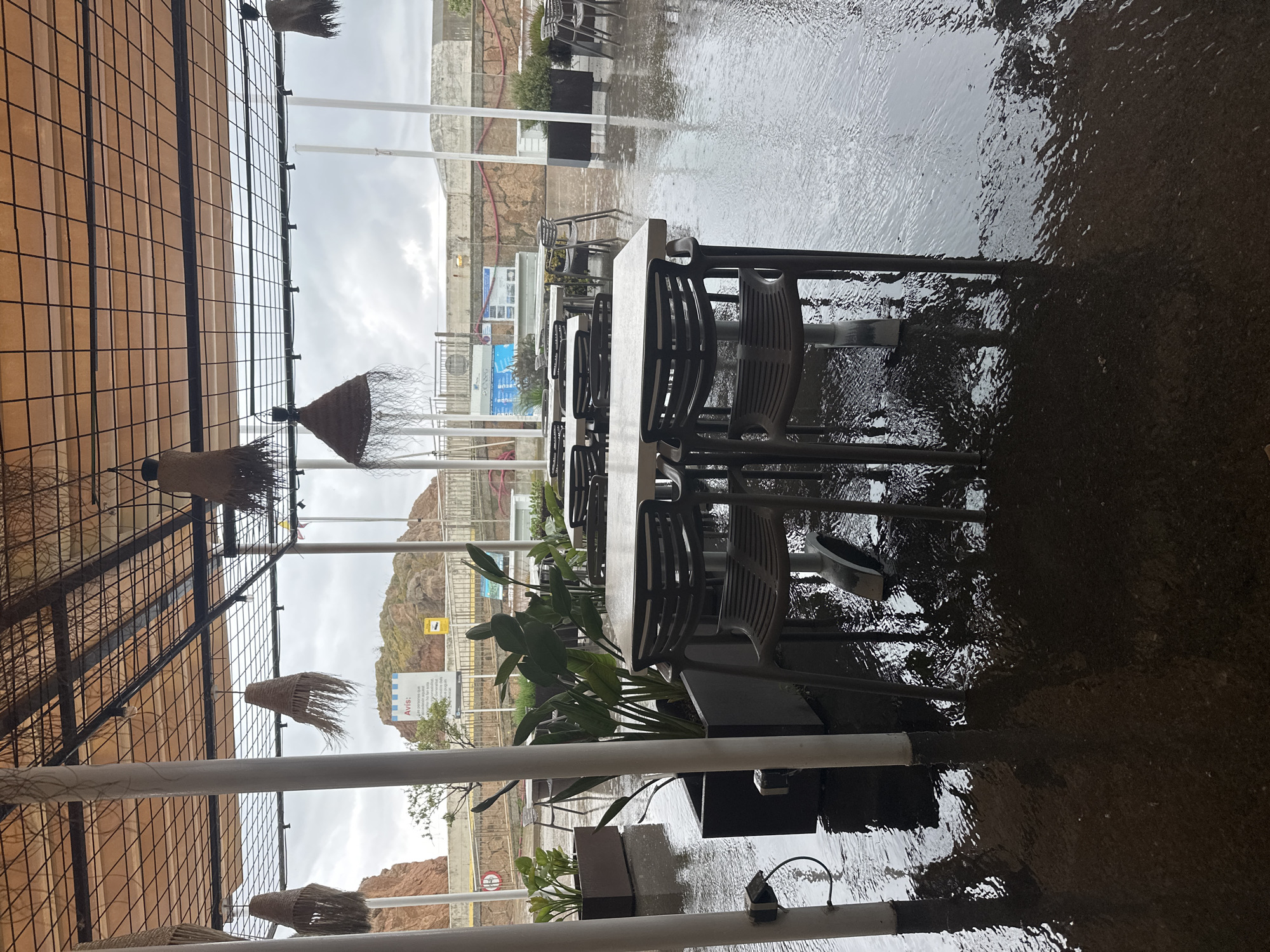 The image depicts an outdoor dining area in Unknown, Spain, likely a restaurant patio. The scene is set on a rainy day, as evidenced by the wet ground and puddles. Tables and chairs, arranged for diners, sit vacant. Several potted plants add greenery to the space. Overhead, a canopy-like structure provides partial shelter, adorned with decorative, possibly straw, lamps. In the background, one can see coastal buildings and cliffs, typical of a seaside setting in Spain. There are no people visible in the image.