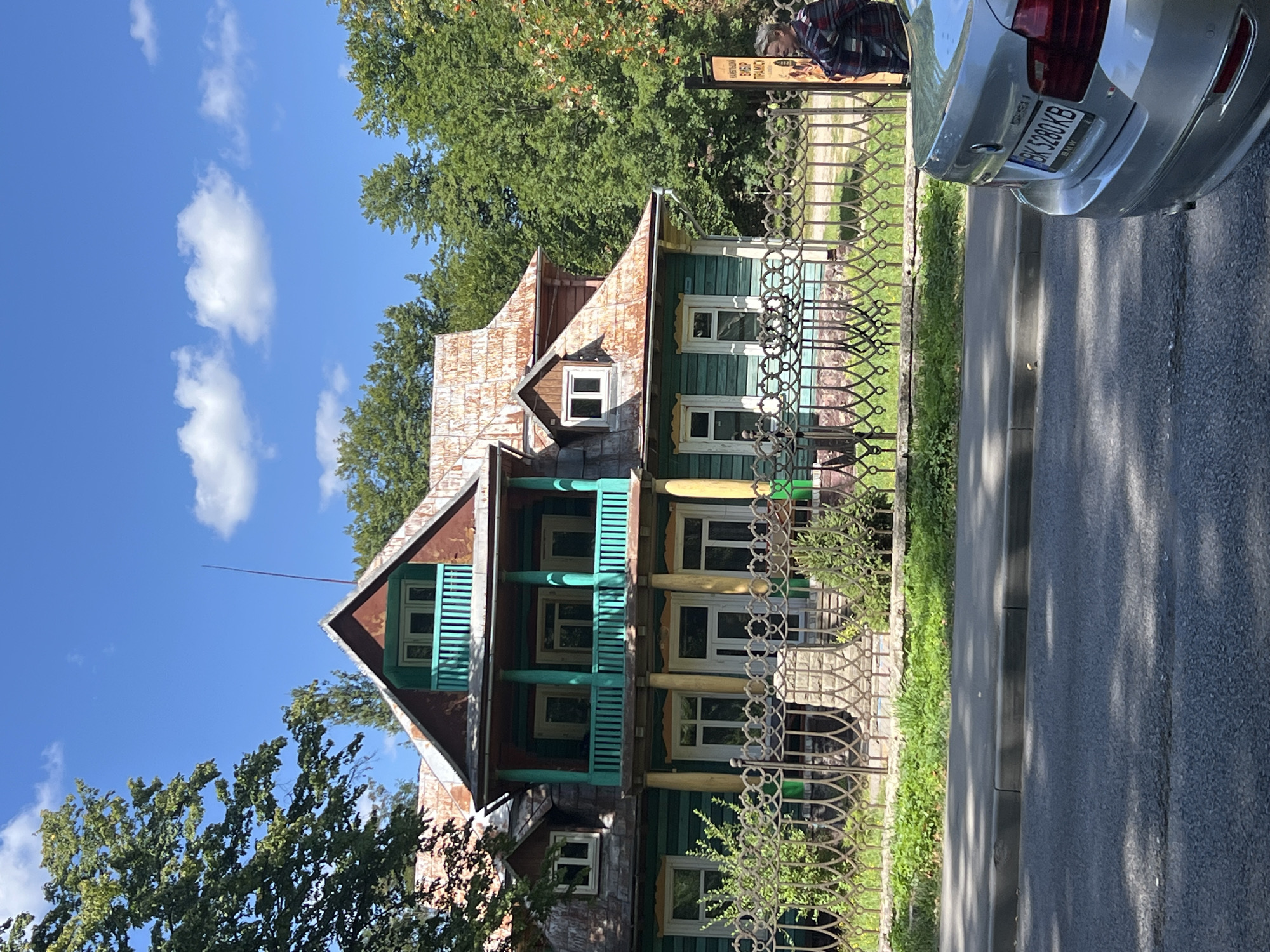 The image depicts a two-story wooden house with green siding and a rusty metal roof. The house features a prominent second-story balcony with teal-colored railings and yellow pillars. Several windows are visible on both floors. A decorative metal fence runs along the front of the property, with a stone-lined garden bed behind it. Lush green trees surround the house, and the sky is bright blue with scattered white clouds. In the foreground, a silver BMW sedan is parked on the right side of the frame. To the right of the house, a person is standing next to a sign, looking towards the house. The setting appears to be a street in the city of Yaremche, Ukraine, known for its wooden architecture.