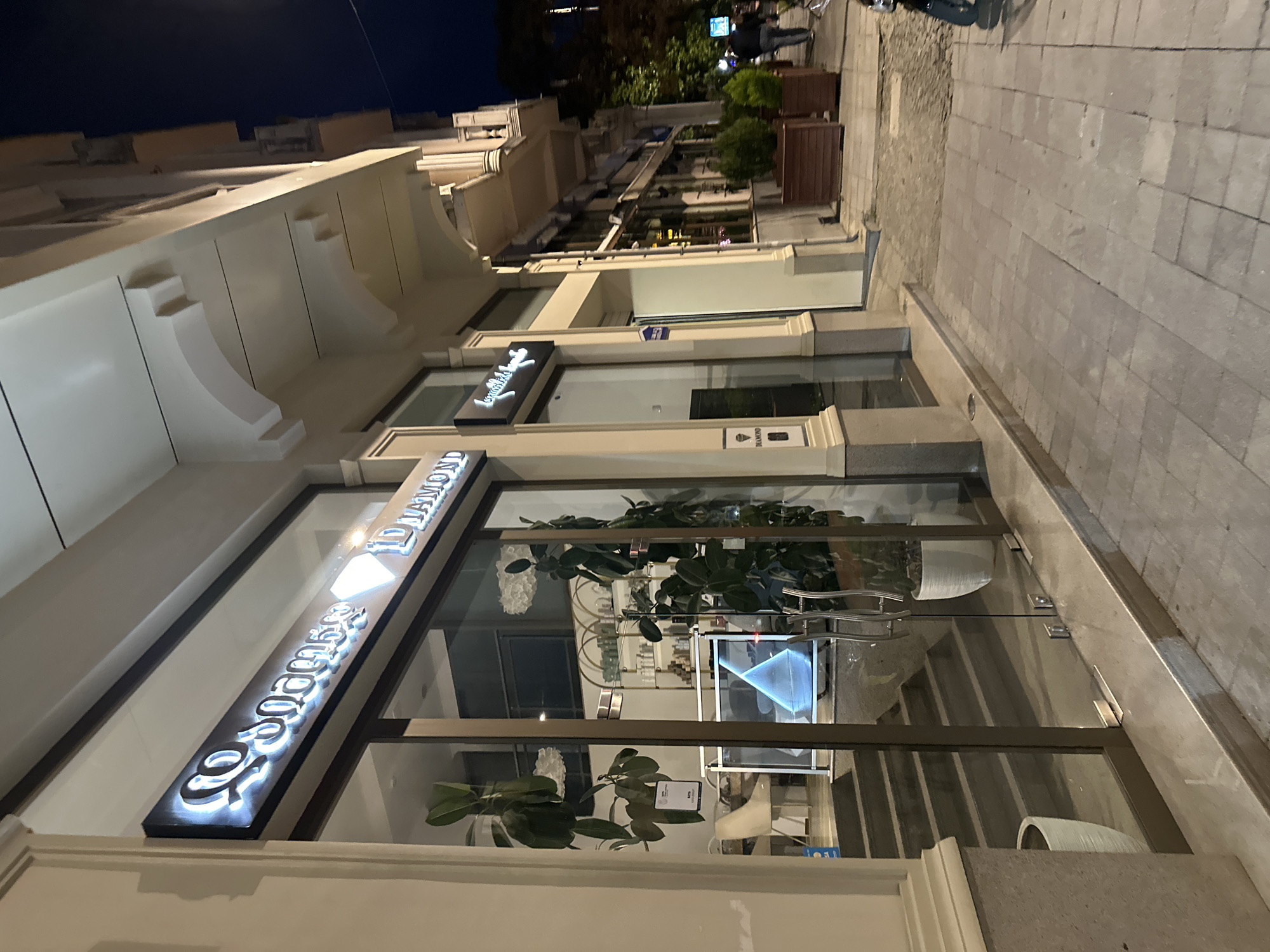 The image shows a street scene in Batumi, Georgia at night. The main subject is a storefront with the illuminated sign "ID DIAMOND" above the entrance. The building has a light-colored facade. The sidewalk is paved with light gray stones. There are large windows in the storefront through which plants and interior details, including a screen or display, are visible. In the distance, other buildings line the street, some with visible balconies and trees. A few people are faintly visible in the background.